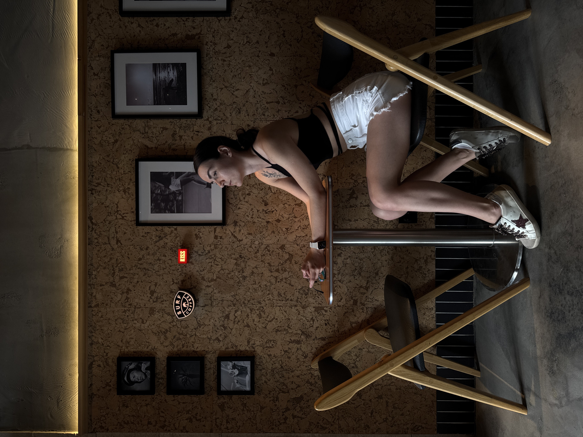 The image shows a young woman in a black tank top and white shorts leaning on a high table in what appears to be an indoor space. She is wearing white sneakers and has her hair pulled back. Her posture suggests she might be looking at something on the table. Two chairs are positioned around the table. The background consists of a wall with a cork-like texture, adorned with framed pictures and a circular "Surf Coffee" sign. A vertical strip of light is visible on the left side of the wall. A small red sign with "KISS" is also visible. The scene is located in Tyumen, Russia.