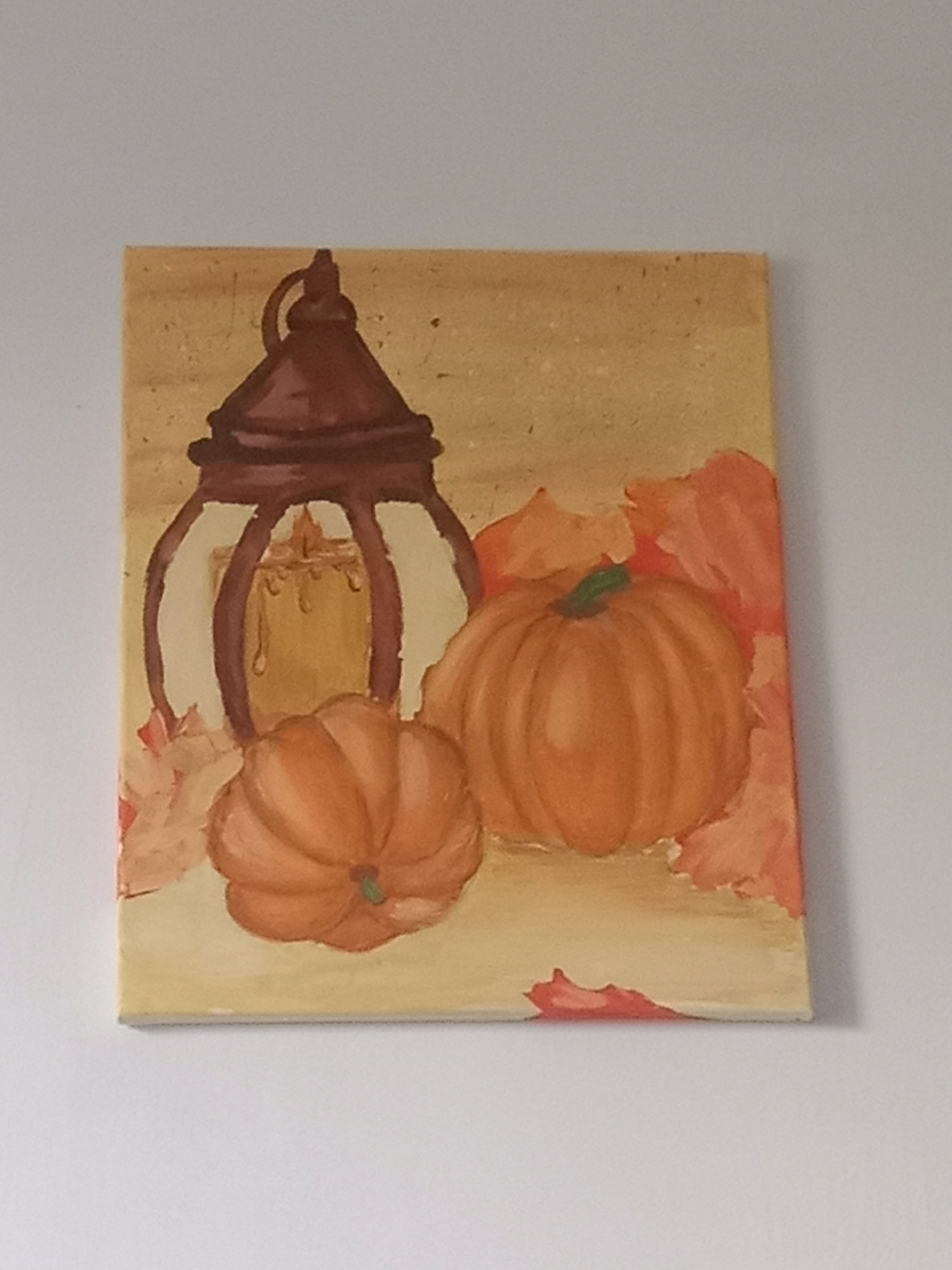 The media file displays a painting of a still life. The subjects are a vintage lantern with a candle inside, two pumpkins, and several autumn leaves. The scene is set against a warm, textured background that suggests a rustic or autumnal ambiance. The painting appears to be done on canvas and is displayed on a white wall. There is no visible text in the image. The lighting is soft, and there are no indications of a specific time of day or weather conditions, though the subject matter evokes a fall theme. The overall mood is serene and cozy, consistent with autumnal decor. The location context of Kielce, Poland, does not provide any specific visual cues within the artwork itself.