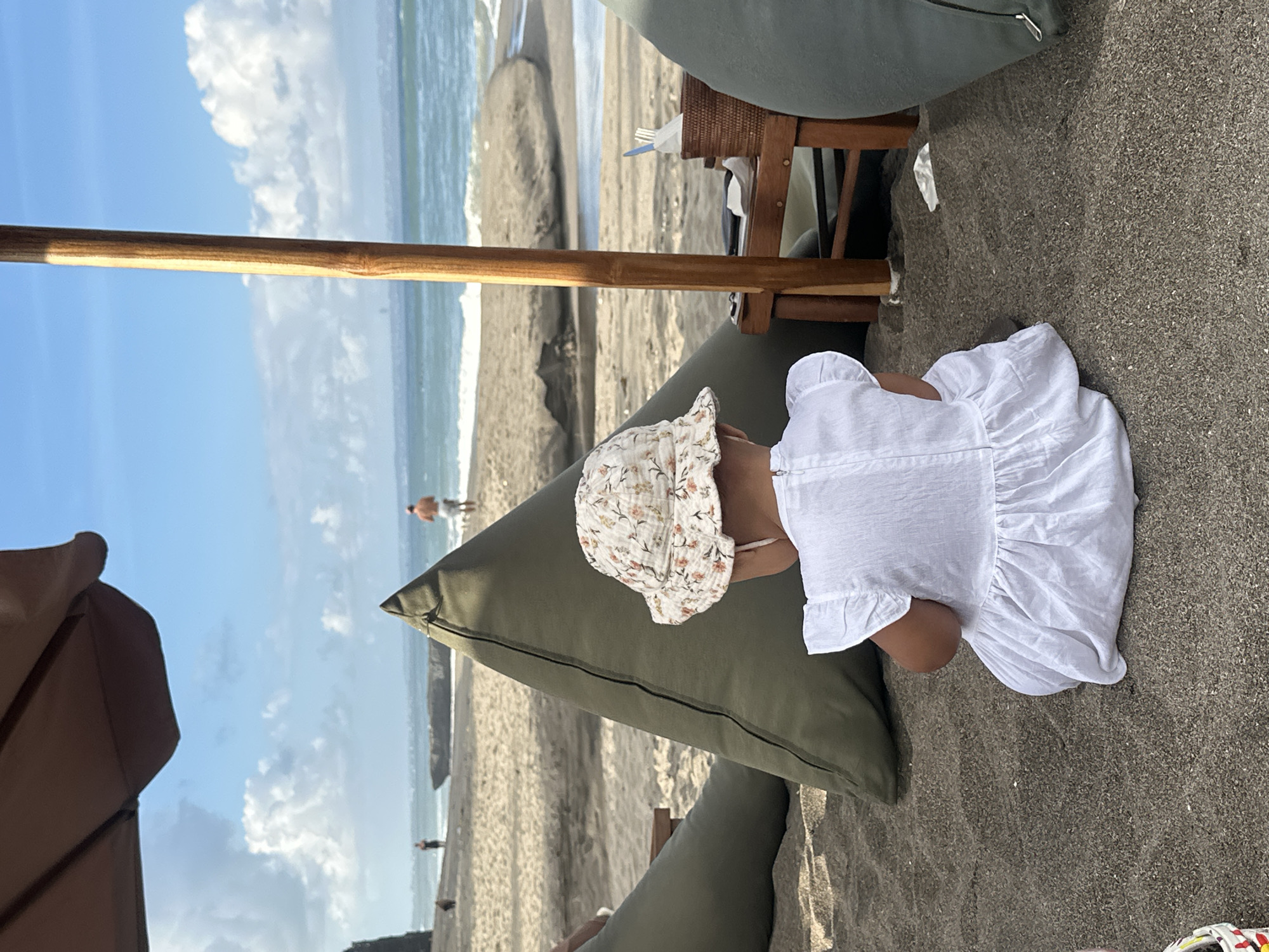 The image depicts a sandy beach in North Kuta, Indonesia. In the foreground, a young child wearing a white dress and a floral-patterned hat is sitting on the dark sand, facing away from the camera. The child is positioned between two large, olive-green beanbag-style seats. Behind the child, a wooden pole supports a brown umbrella, partially visible at the top of the frame. To the right of the pole, a small wooden table holds a woven basket with cutlery and napkins. The middle ground shows the beach with more beanbags, and further back, people can be seen walking near the ocean. The ocean has moderate waves, and the sky is blue with scattered white clouds. The overall scene suggests a relaxing day at a beachside establishment.