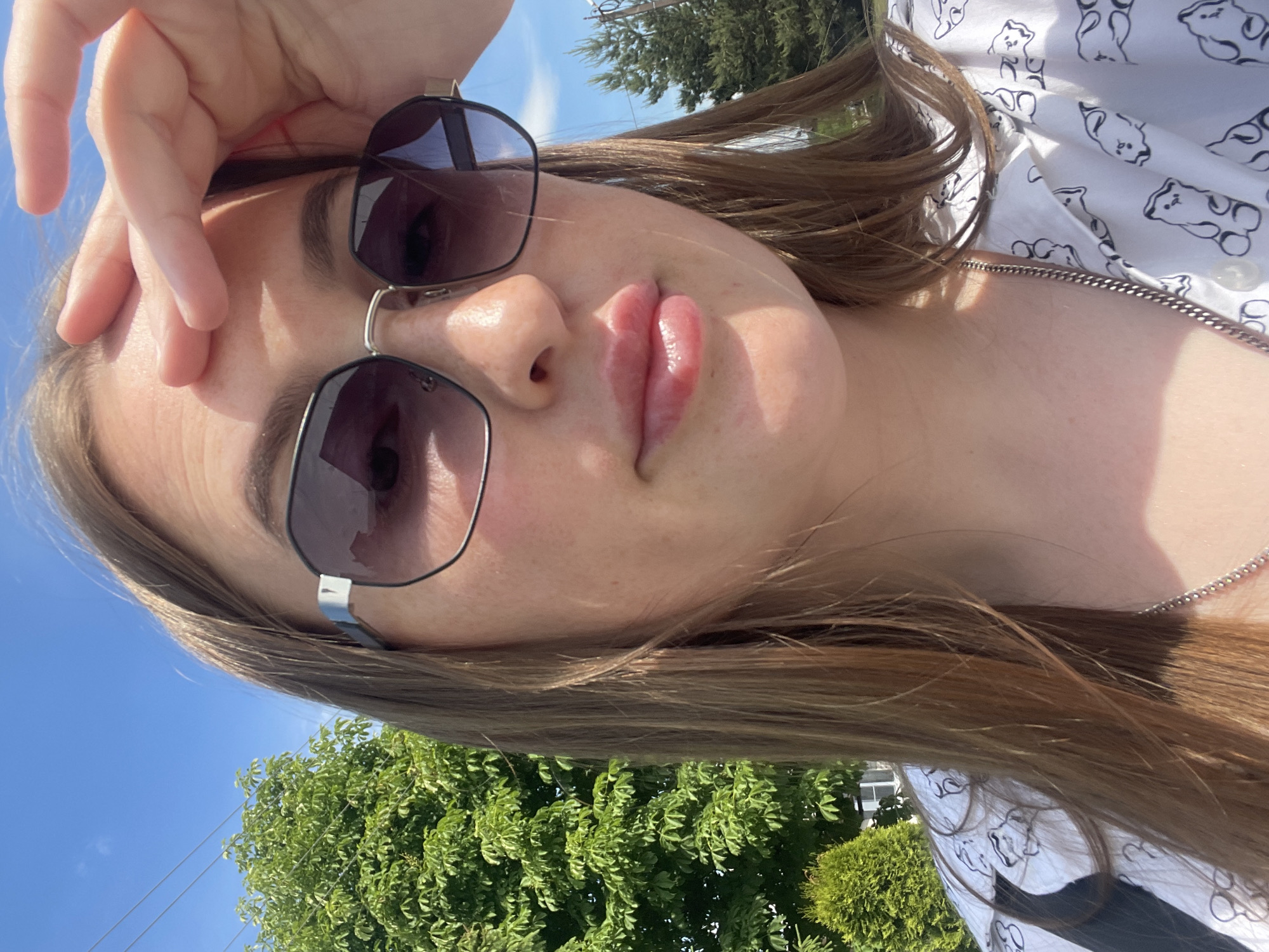 A young adult female is captured in a sunlit outdoor selfie, her right hand (viewer's perspective) raised with fingers resting on her forehead, appearing to shield her eyes from direct sunlight. She has long, straight, light brown hair and is wearing dark, hexagonal-framed sunglasses. Her lips are slightly pursed, and freckles are visible on her nose and cheeks. She is dressed in a white collared shirt adorned with a repeating pattern of black outline teddy bears, and a silver-colored chain necklace is visible around her neck.
The background consists of a clear blue sky with thin power lines stretching across the upper left. Lush green deciduous trees are prominent on the left side of the frame, with a smaller, conical evergreen shrub visible at the lower left. A partial view of a dark-needled coniferous tree is seen on the right, behind her shoulder, along with what appears to be a segment of a utility pole. A sliver of a white building is visible in the distant background on the left. The scene is brightly illuminated by direct sunlight, casting highlights and shadows. This image was captured in Сырково, Russia.