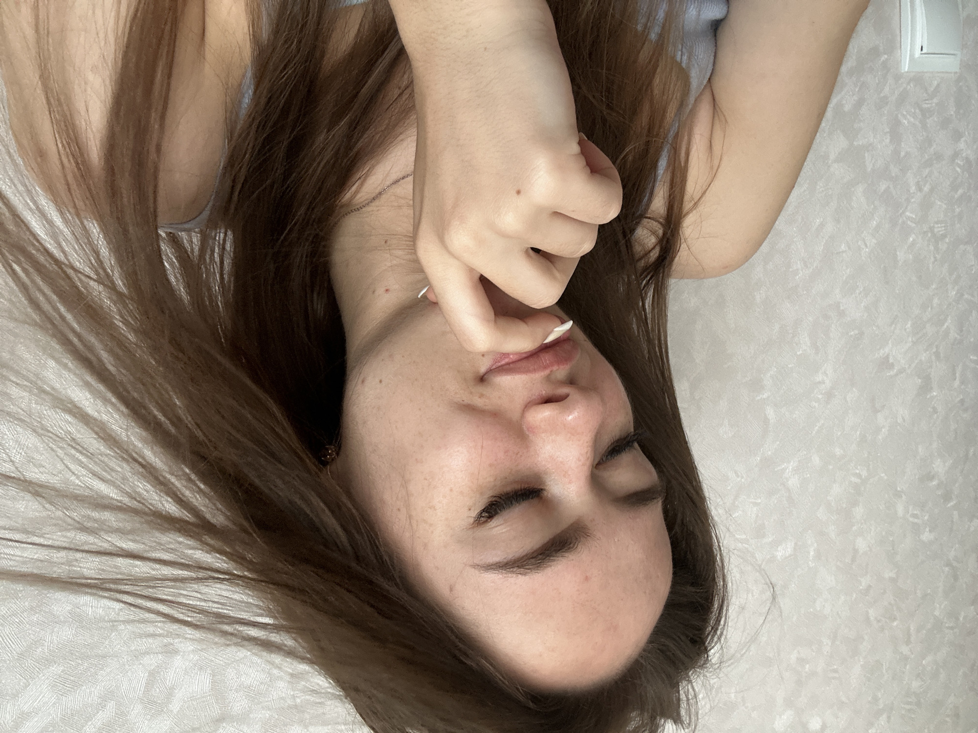 An overhead, upside-down close-up captures a young person, likely female, lying down with their eyes closed. Long, straight brown hair fans out around their head, partially obscuring a light blue or grey top. One hand rests near their chin and mouth, fingers slightly curled, revealing white fingernail polish and a small mole on the back of the hand. A faint necklace chain is visible around the neck. The subject's face shows several small freckles.
The background consists of a light-colored, textured surface, possibly a wall or bed sheet. In the upper right corner, a white light switch or electrical outlet is partially visible against this background. The lighting is soft and even, suggesting an indoor setting. There is no visible text in the image. The overall mood conveyed is one of tranquility or contemplation due to the closed eyes and relaxed pose.