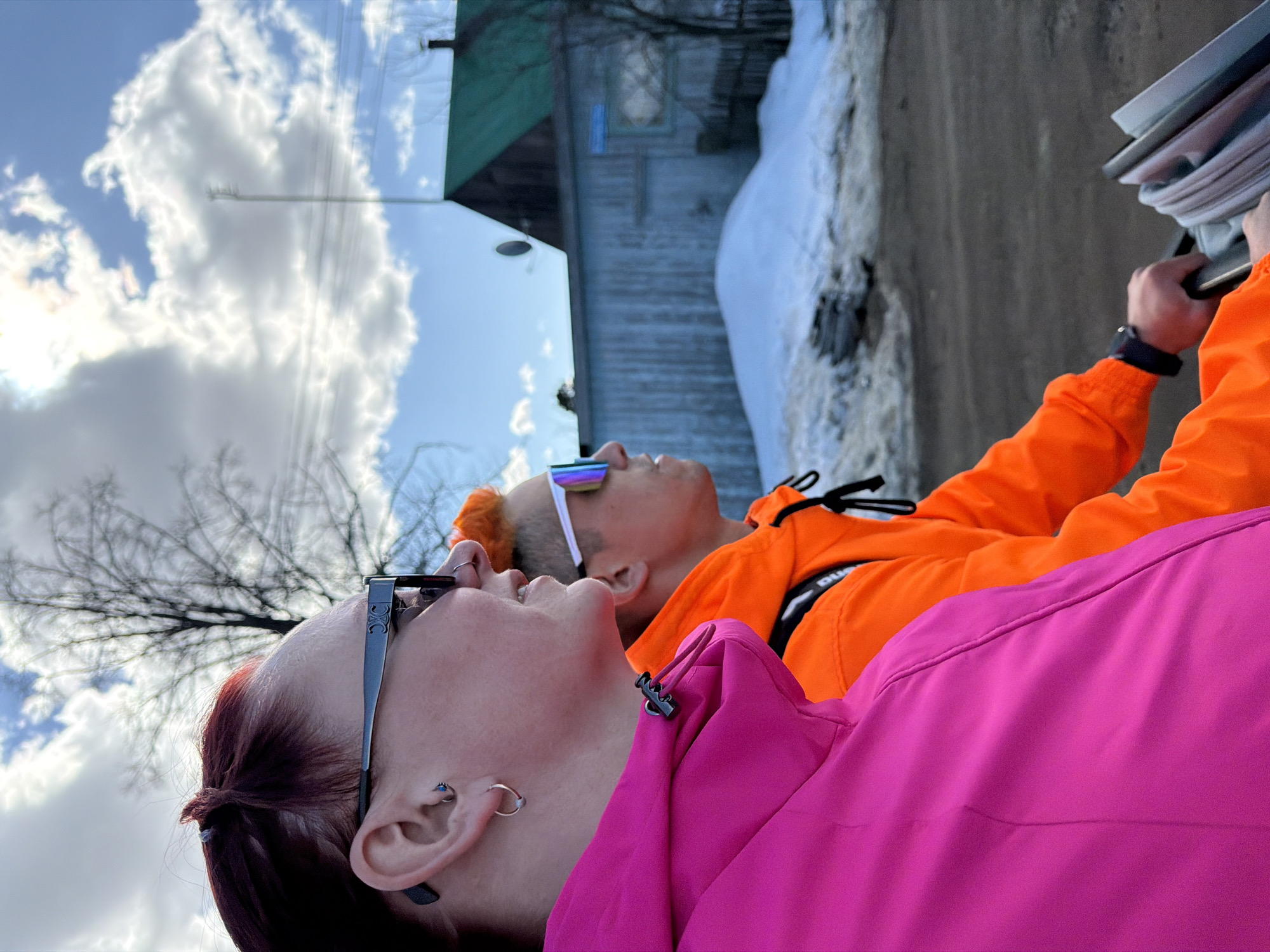 Two people are looking up at the sky in Chaikovsky, Russia. One person is wearing a pink jacket and glasses, and the other person is wearing an orange jacket and colorful sunglasses. A building and some snow are visible in the background. The sky is blue with white clouds.