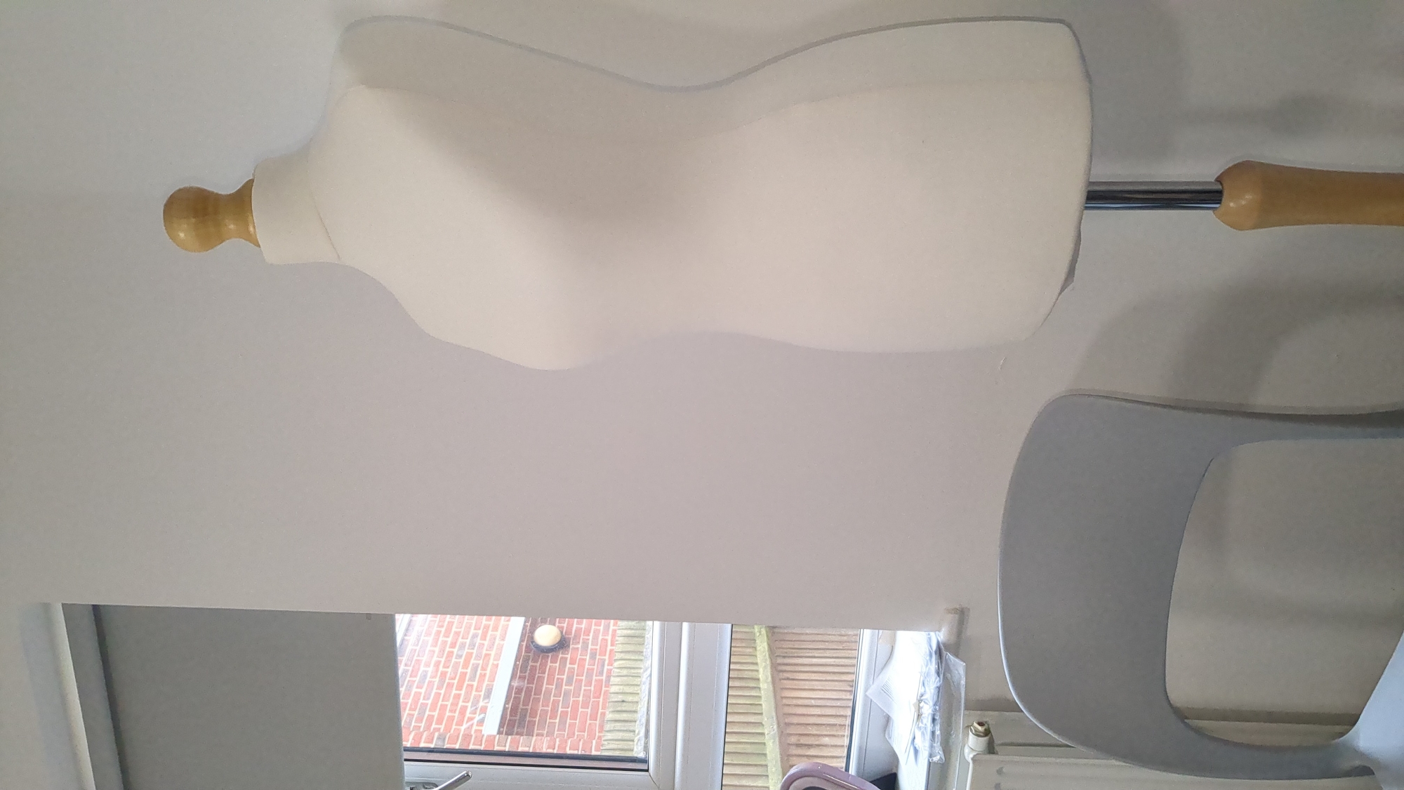 A dressmaker's dummy, with a white fabric-covered torso and a wooden neck and stem, is partially visible in an interior setting. A window in the background reveals an exterior view of a brick building and slatted blinds, indicative of residential architecture. A curved, light-grey object, possibly part of a chair or stand, is also present in the foreground. The location can be inferred to be in Epsom and Ewell, United Kingdom, based on the prompt's request.