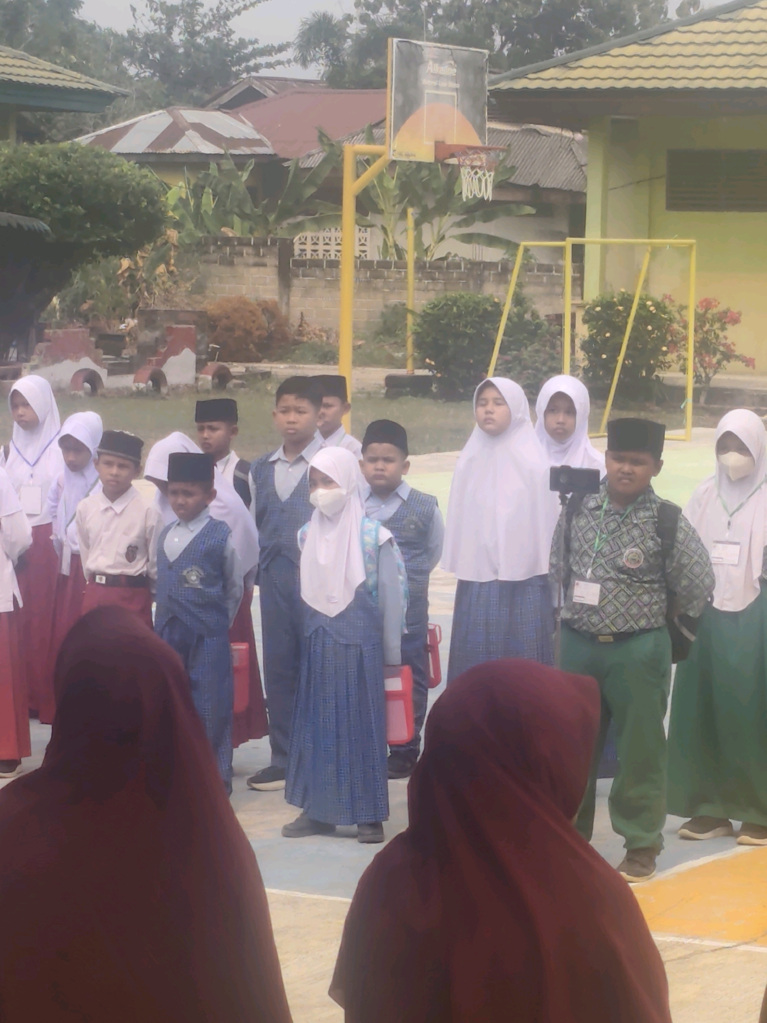 In Pematang Reba, Indonesia, on the grounds of a school, a group of students stands assembled. The students, some wearing hijabs and sarongs, are dressed in various uniforms. The backdrop includes buildings with pitched roofs, a basketball hoop, and a chain-link fence, suggesting an outdoor educational setting. The image appears to be taken during daylight, with a slightly hazy sky.