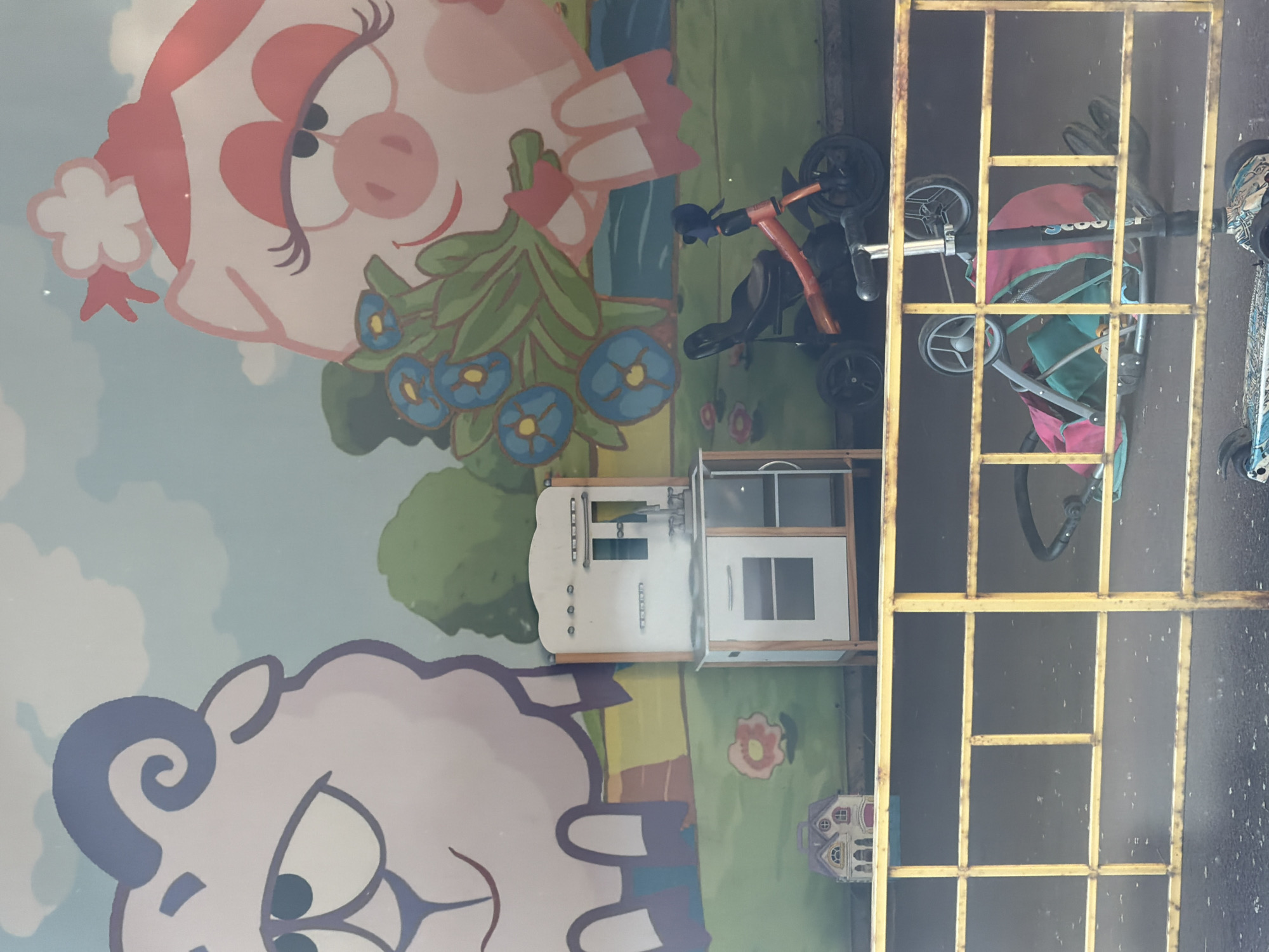 A mural of cartoon characters, a sheep and a pig, is visible on a wall in what appears to be an outdoor public space. In front of the mural, there are several children's toys. A miniature kitchen set with a white front and wooden frame stands against the wall. To the right of the kitchen set, a red and black tricycle is parked. In the foreground, a yellow metal fence separates the viewer from a stroller with a pink and turquoise fabric, and parts of a scooter are visible on the ground. The ground appears to be asphalt, with some wet patches. The overall scene suggests a play area or an outdoor entertainment zone for children, potentially located in the city of Krasnoyarsk, Russia, given the provided context. There are no people visible in the image.