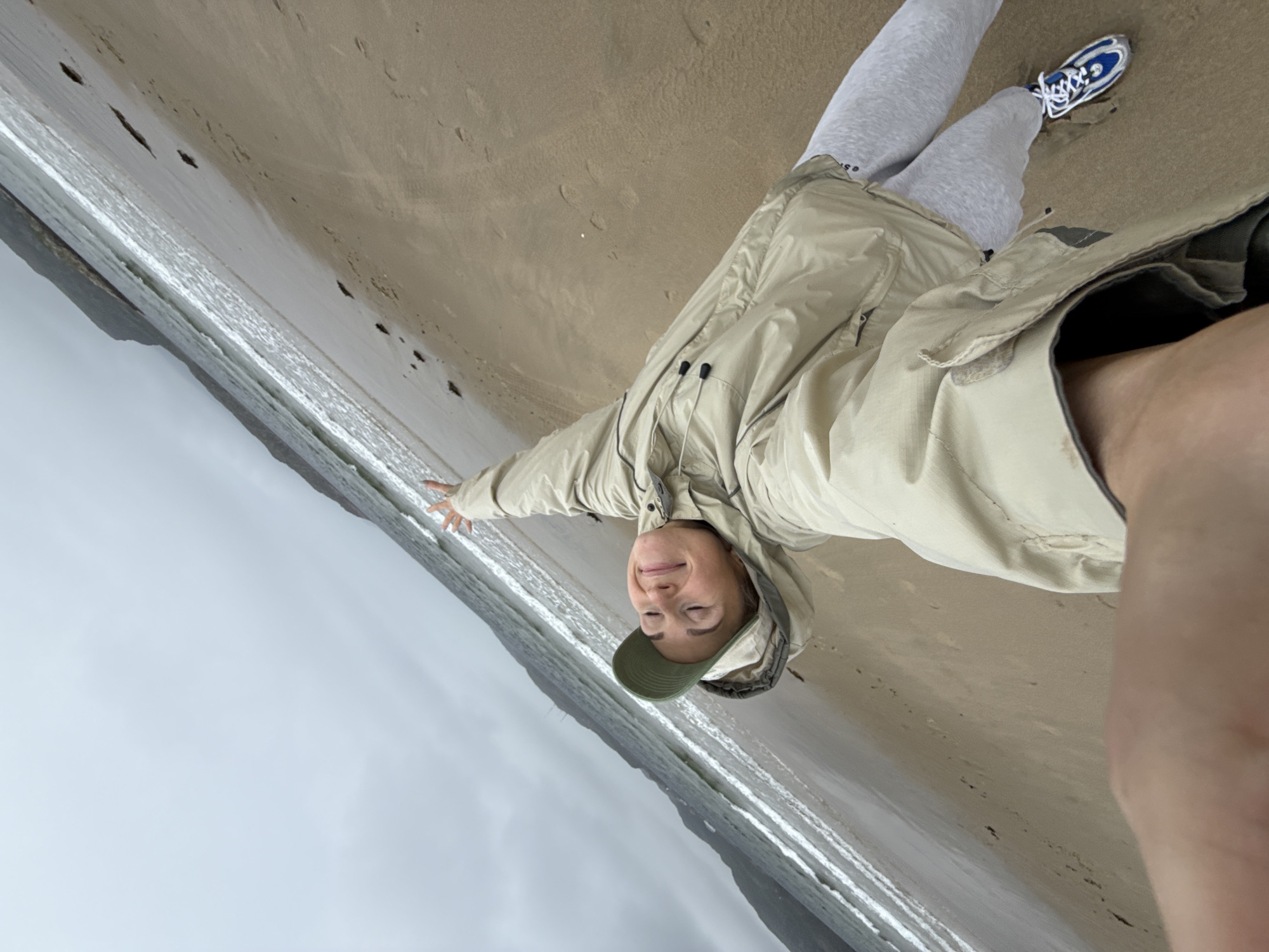 A woman with blonde hair and closed eyes is lying on a sandy beach, holding a camera to take a selfie. She is wearing a light brown jacket, a green baseball cap, and light gray sweatpants. Her right arm is raised in the air. The beach is wet from the tide, and there are small waves rolling in from the ocean. In the background, there are mountains under a cloudy sky. The image is taken at an angle, giving a sense of movement. This scene appears to be the shore of the Barents Sea in Teriberka, Russia.