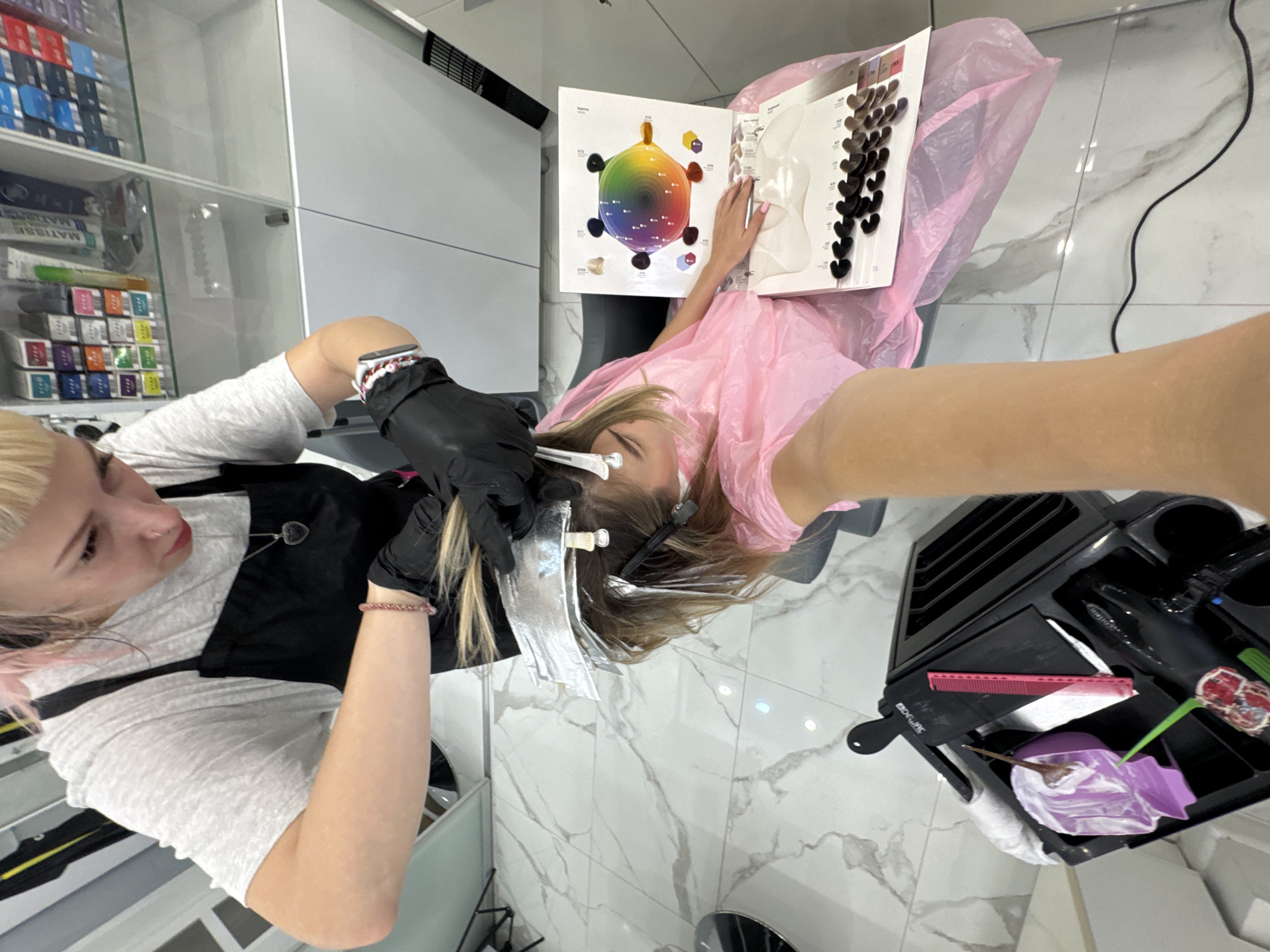An overhead, medium shot captures a hair stylist in a black apron applying hair color to a client in a pink smock within a brightly lit salon. The stylist, wearing black gloves, works on the client's blonde hair, which is sectioned with foil and clips. A color wheel and swatches are displayed on a nearby surface, aiding in the color selection process. In the foreground, a black hair care cart holds various tools, including combs, brushes, and a hair dryer. The salon features a marbled floor and reflective surfaces, indicating a modern and clean environment. The image appears to have been taken from the perspective of the person taking the selfie.