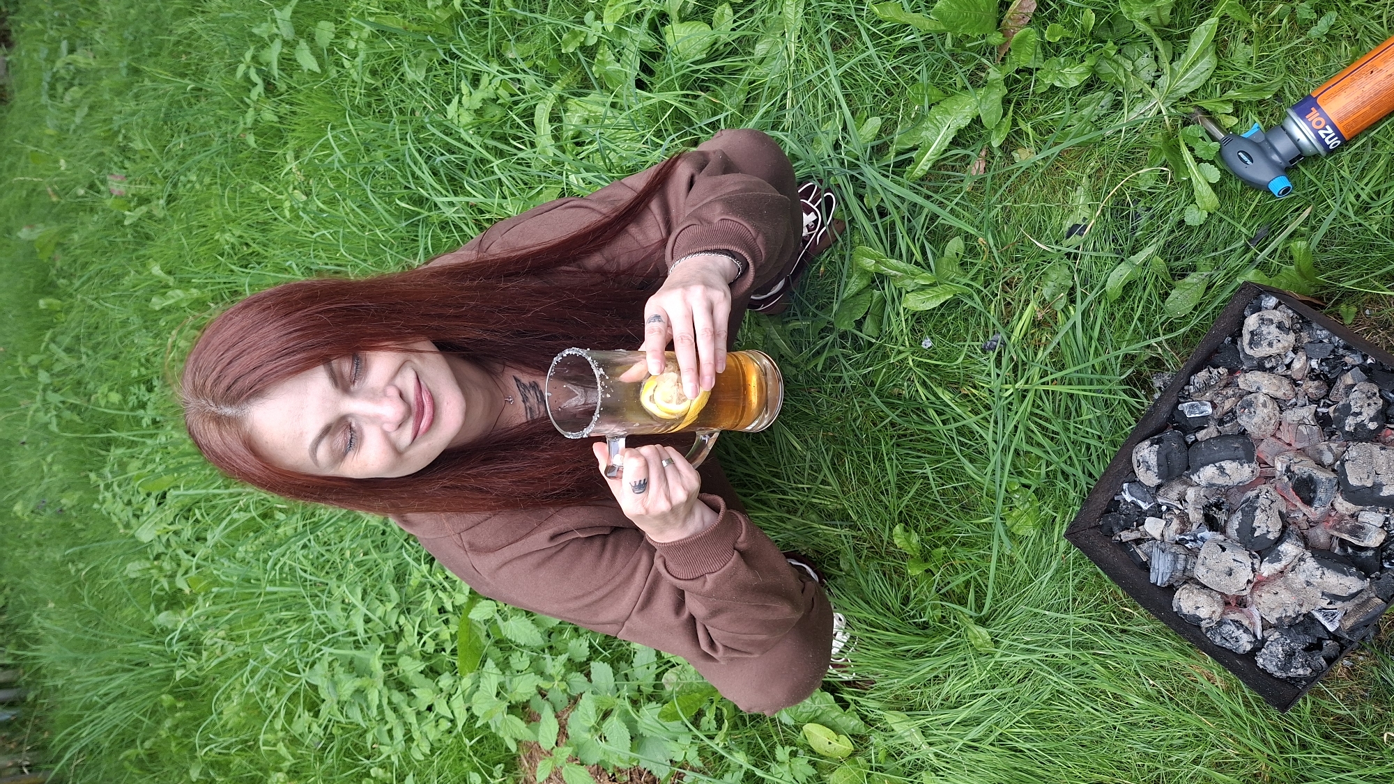 A woman with long, auburn hair sits on a grassy lawn, holding a tall glass of a light-colored beverage with lemon slices. She is wearing a brown hoodie and pants. Her eyes are closed, and she has a slight smile, suggesting a relaxed and pleasant mood. The setting appears to be outdoors, likely a backyard or park, given the lush green grass and surrounding vegetation.
To her right, there is a barbecue grill filled with glowing coals, indicating that a barbecue or grilling activity has recently taken place or is about to begin. Next to the grill is a small gas torch with an orange body and a blue nozzle, labeled with the word "ORZOIL".
The lighting and the vivid green of the grass suggest it is daytime, likely during the warmer months. The overall atmosphere is casual and relaxed, with the woman enjoying a drink in a natural, outdoor setting. The visible text "ORZOIL" is on the gas torch. The location context is Забалоцкі сельскі Савет, Belarus.