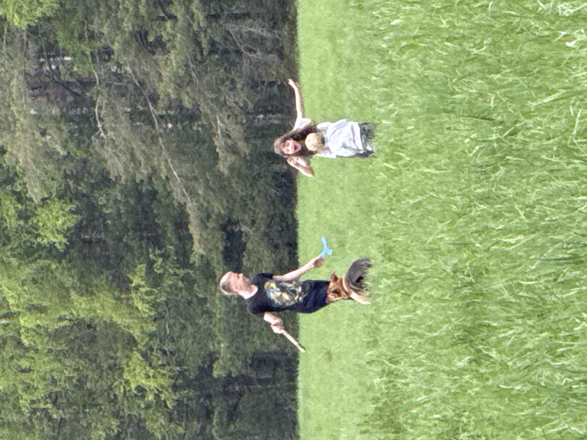 The image depicts a grassy field adjacent to a wooded area. A man wearing a black t-shirt and black shorts is holding a blue object in his right hand and a stick in his left. A woman with long dark hair and a child are standing together, with the woman's right arm raised. The location is Малая Рукавицкая, Russia.
