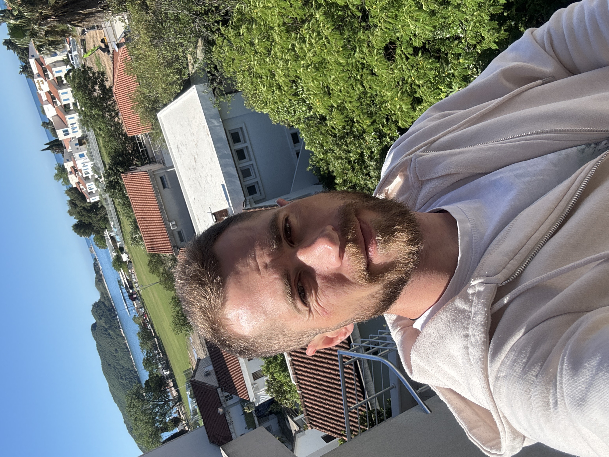 The image is a selfie of a man with short brown hair and a beard, wearing a light-colored zip-up hoodie over a white t-shirt. He is standing outside, overlooking a scenic landscape. In the background is a view of the city of Budva, Montenegro. There are multiple buildings with orange tile roofs and white walls. A strip of green grass runs along the coast. Further in the background is a body of water, possibly the Adriatic Sea. A green, tree-covered peninsula juts into the water. The sky is blue. The man appears to be on a balcony or terrace, based on the presence of a metal railing.