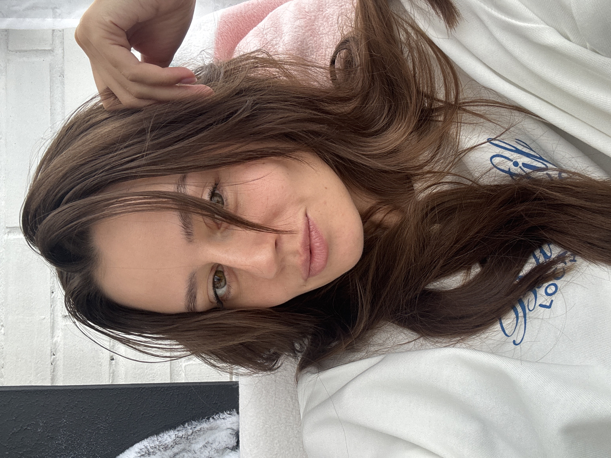 A woman with long, wavy brown hair is reclining, with her head and shoulders visible. She has light skin, light brown eyes, and pink lips. Her left hand is raised, touching her hair. She is wearing a pink top and lying on a white surface with a blue design. The background includes a white brick wall and a black and white object. The location is Krasnoyarsk, Russia.