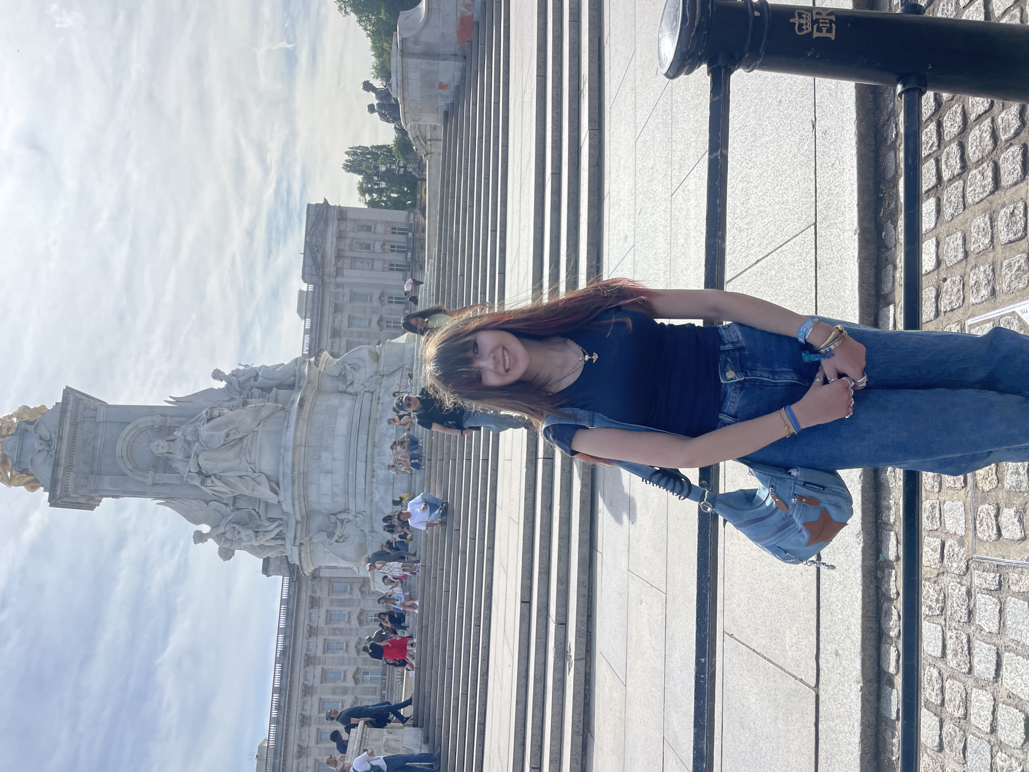 A young woman with long, dark hair and bangs, wearing a dark blue t-shirt and blue jeans, poses for a photo outdoors. She is smiling and has a blue denim bag slung over her left shoulder. In the background, a large white monument topped with a golden figure stands prominently, with a grand building behind it. Several other people are visible in the background, some sitting on stairs and others walking. The scene appears to be Buckingham Palace in London, United Kingdom, given the architecture of the palace and the Victoria Memorial in front of it. The lighting suggests it is daytime, with some clouds in the sky.