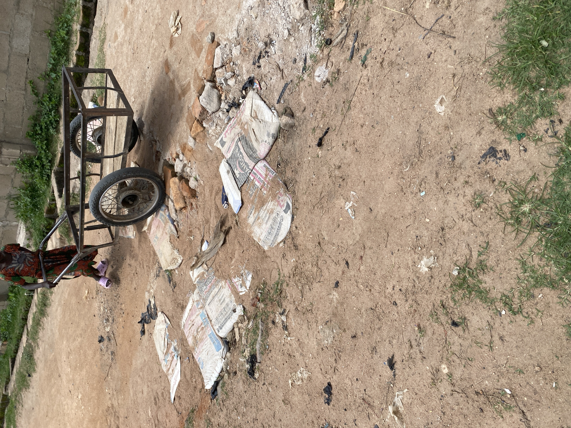 In Jauro Yinu, Nigeria, a person in a vibrantly colored garment pushes a two-wheeled cart made of metal. The cart is empty. In the foreground and scattered across the dusty ground are several large, white bags, some appearing to be cement bags with branding like "DANGOTE" and "MASTER CEMENT" visible. Various pieces of debris, including plastic bags, rocks, and twigs, are strewn about the environment. The ground is composed of dry dirt with patches of sparse green grass. In the background, a low concrete wall and some greenery suggest the edge of a settlement.