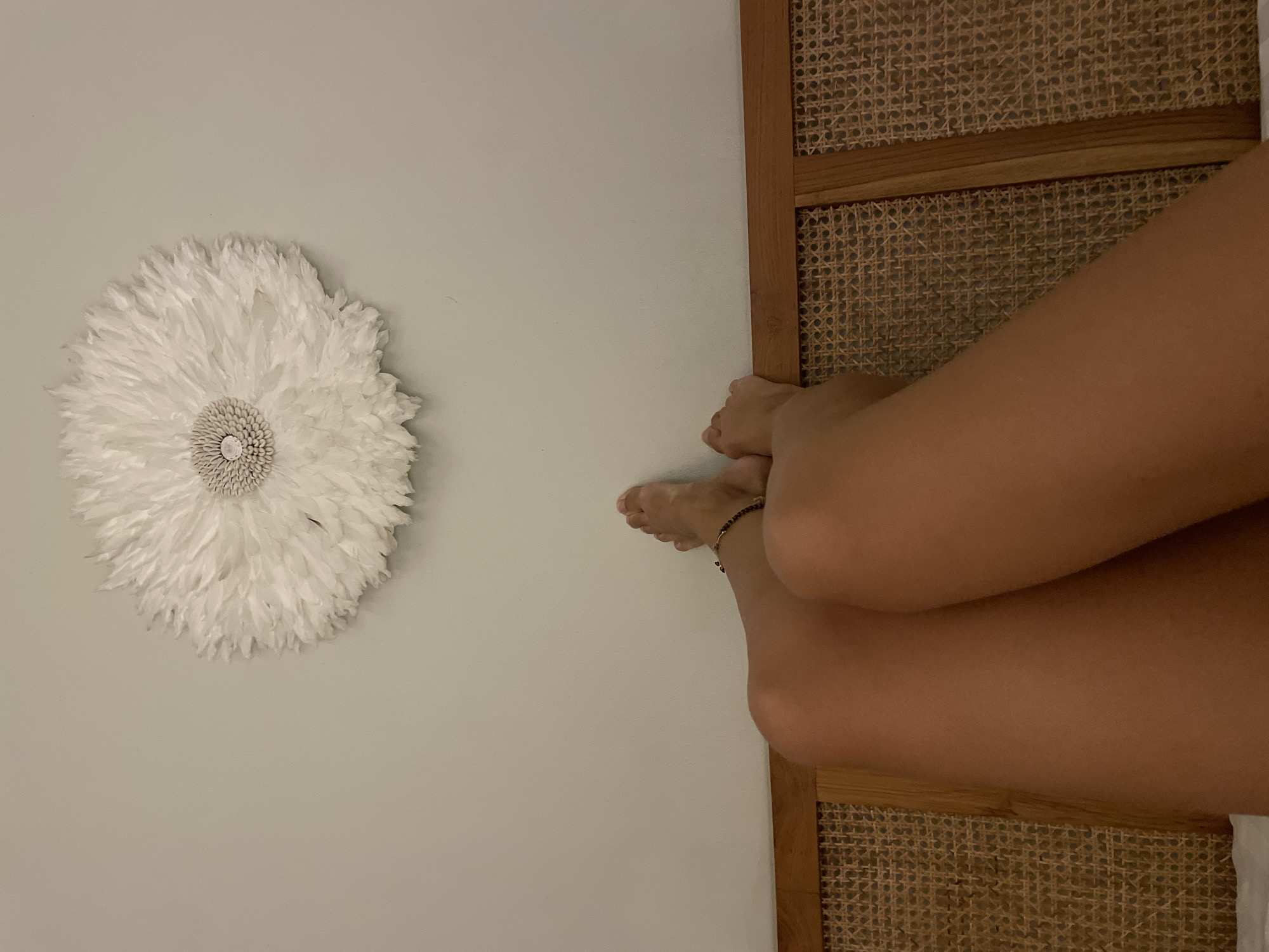 The image shows a room with a wall adorned with a white decorative feathered item. Adjacent to the wall is a rattan headboard, with a person's legs and feet positioned near it. The person is wearing a bracelet on their ankle. The location is Unknown, Unknown.