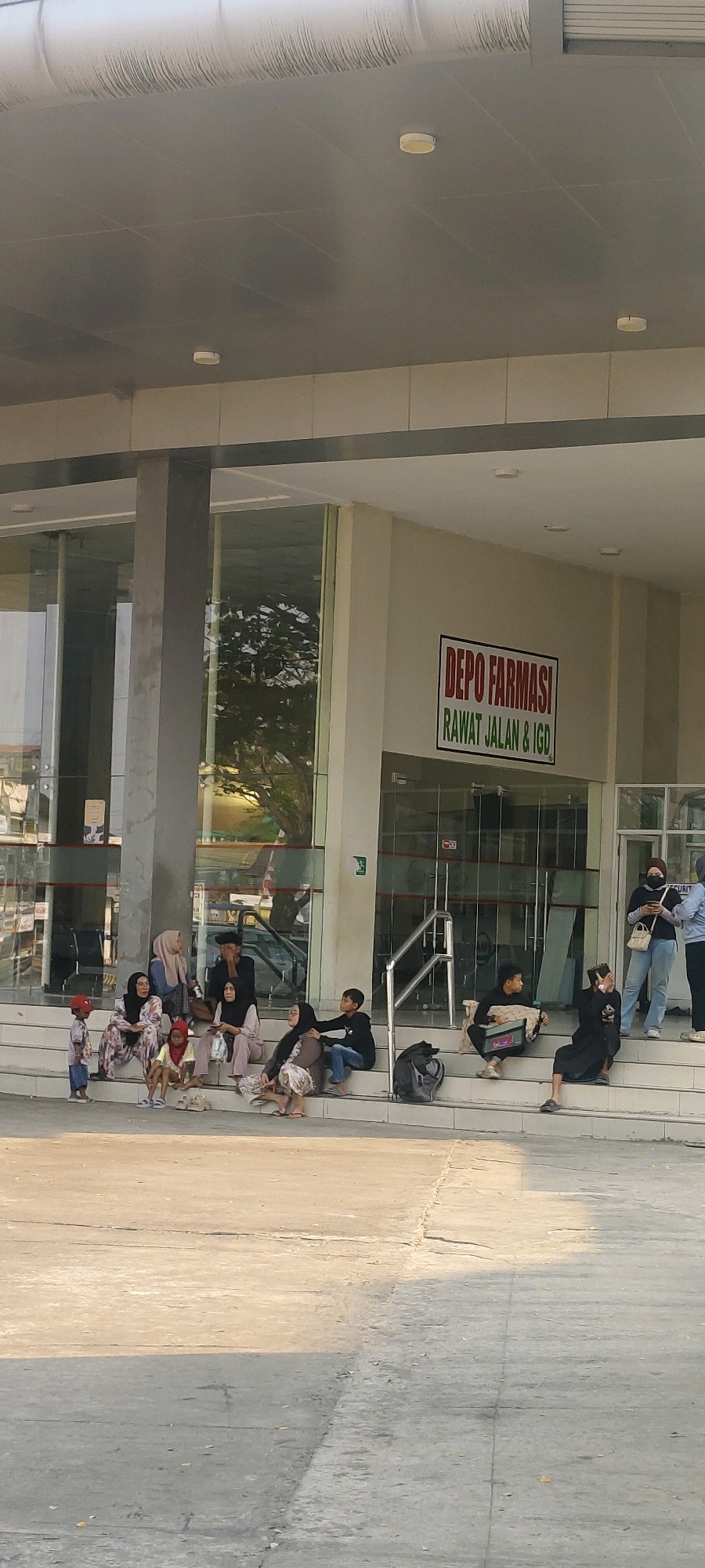 A group of people are seated on concrete steps outside a building with large glass windows. A sign above the entrance reads "DEPO FARMASI RAWAT JALAN & IGD". The building appears to be a pharmacy or clinic. The people are dressed casually, and some are wearing headscarves. A child in a red shirt and blue pants stands on the ground to the left of the steps. The scene suggests a waiting area or public space. The location is identified as Banjarmasin, Indonesia.