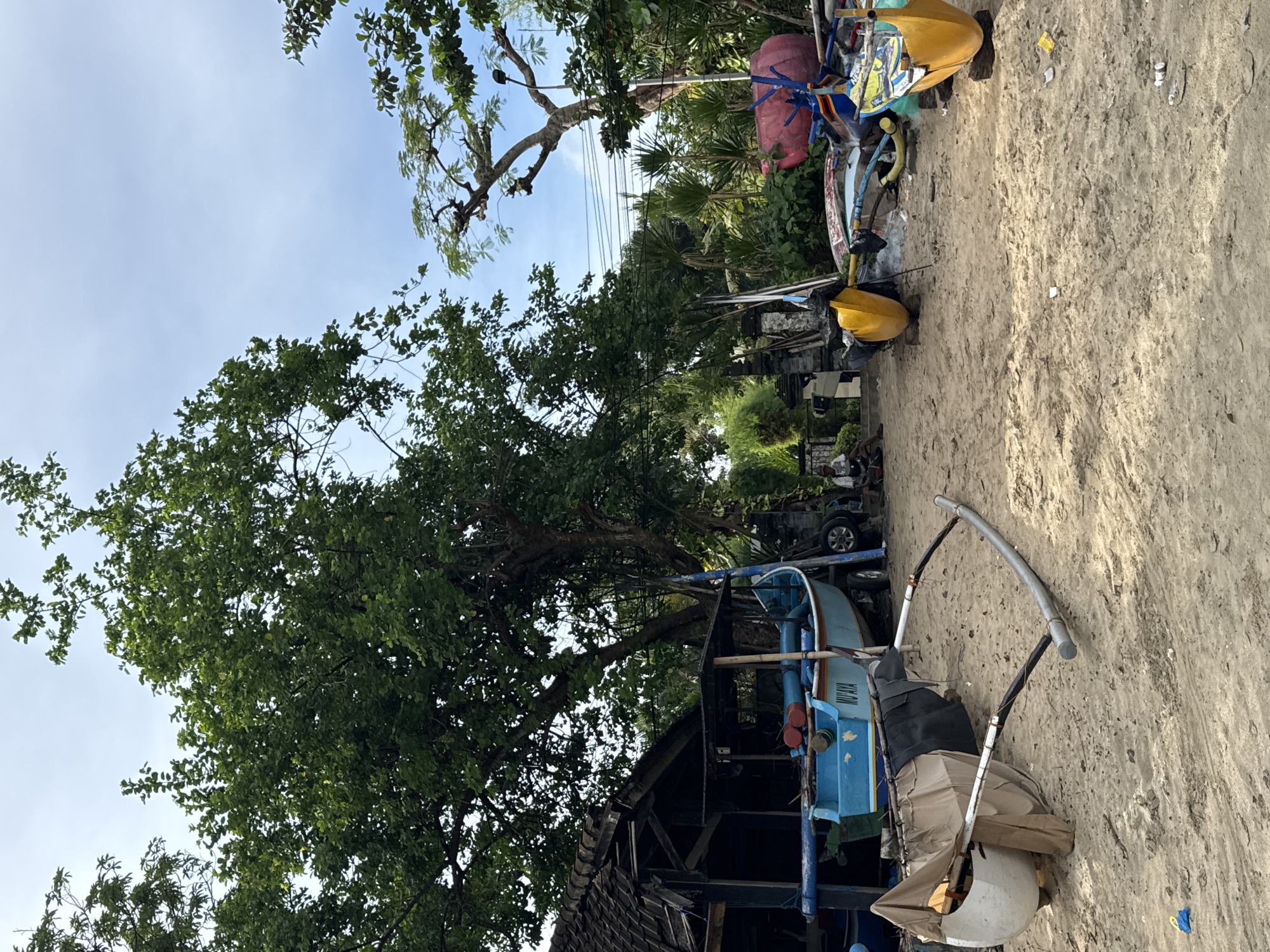 The scene is a beach in Kuta Selatan, Indonesia. Sand dominates the foreground and leads to a cluttered area with various objects. A blue boat rests near a dilapidated wooden structure with a damaged roof. Trees with dense green foliage are abundant, partially obscuring the sky. In the background, there are more trees, a car, and some manmade structures. Debris is visible on the sand. The sky is a light blue.