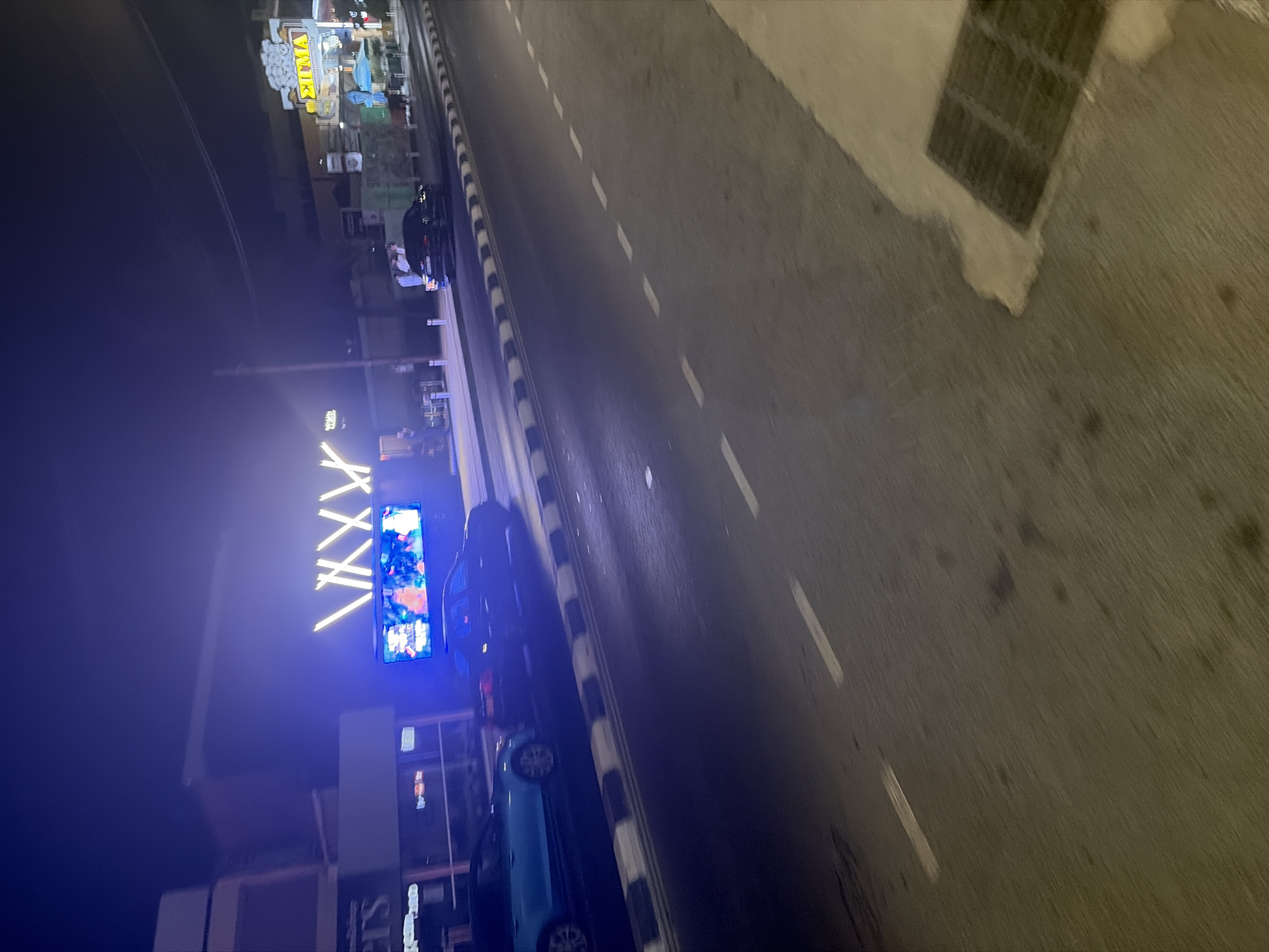 The image depicts a street scene at night in Agios Athanasios Municipality, Cyprus. A bright blue neon sign with a "V" pattern is prominently displayed on a building facade, along with a digital screen showing animated images, likely advertising a venue. In front of this building, several individuals are standing on the sidewalk, illuminated by the neon lights. A dark-colored sedan is parked on the street, and a light blue sedan is also visible on the road. To the right, another building features a yellow illuminated sign with Greek lettering, "ΣΙΓΜΑ," suggesting a business or establishment. The street is paved, with a distinct black and white striped curb. The environment is dark, with artificial lighting from the signs and streetlights illuminating the scene. A storm drain is visible in the lower right corner of the image.