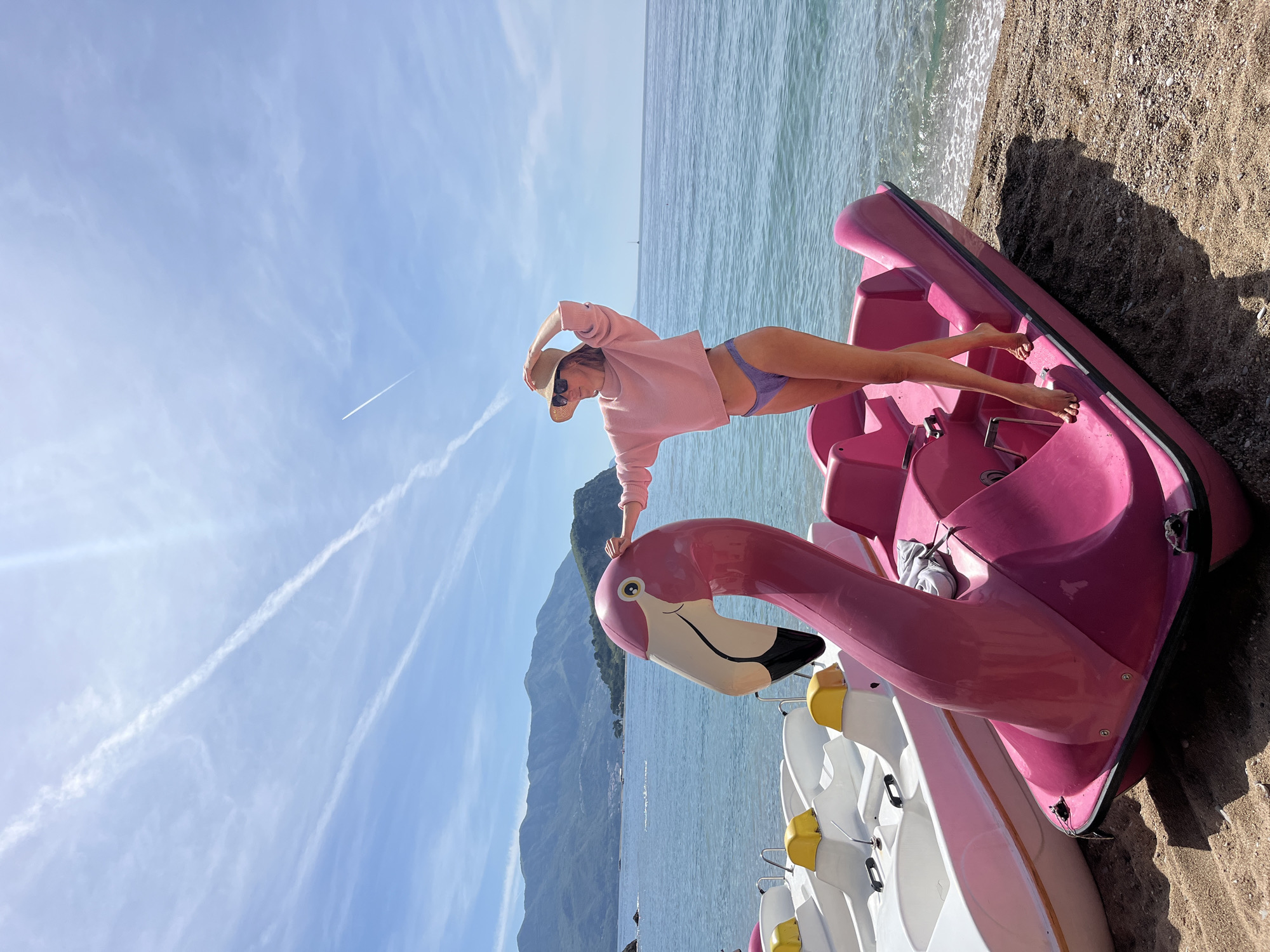 A woman stands on a flamingo paddle boat on a beach in Budva, Montenegro. She is wearing a pink sweater, lavender bikini bottoms, and a straw hat. She has sunglasses on and her left hand on her head. The flamingo boat is pink and there are white paddle boats behind it. The beach is made of sand and the water is turquoise. There are mountains in the background and a blue sky with contrails.