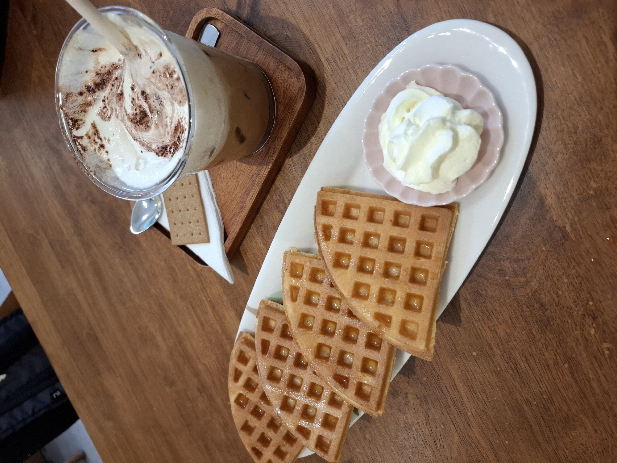 A close-up, high-angle shot shows a dessert setting on a wooden table. In the foreground, a white, oval-shaped plate holds four quarter-circle waffles, each with a golden-brown, crispy exterior and a syrup-like glaze. To the right of the waffles is a small, pink, scalloped dish containing a swirl of white whipped cream. In the background, a clear plastic cup contains a cold coffee drink with ice cubes and a generous topping of whipped cream swirled with cocoa powder. A light-colored straw protrudes from the drink. A single square biscuit and a small silver spoon rest on a white napkin. The wooden table has a warm, brown grain. The scene is devoid of people, indicating a solo meal or a moment captured before or after someone enjoyed the food. There are no visible contextual clues to pinpoint the location to Nha Trang, Vietnam.