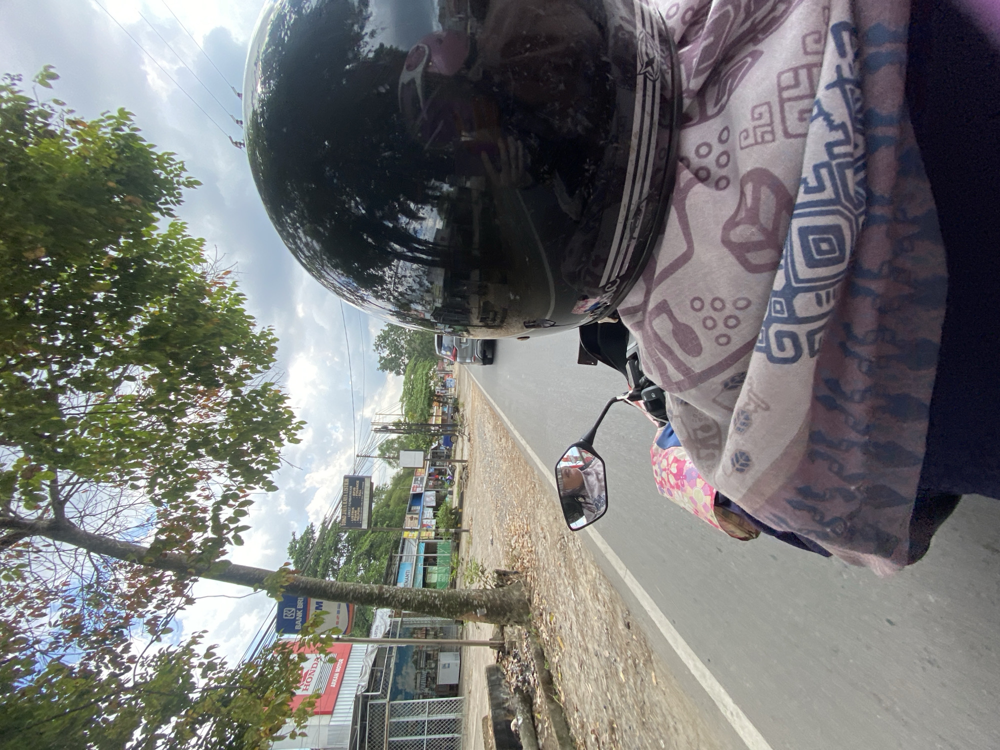 The image captures a first-person perspective of someone riding a motorcycle on a paved road in Banjarbaru, Indonesia. The rider is wearing a black helmet and a patterned scarf. In the rearview mirror, a person wearing glasses and a headscarf is visible. Along the left side of the road, there are buildings, trees, and a sidewalk. Signs for "BANK BRI" and "HONDA" are visible on some of the buildings. The sky is cloudy. The road ahead shows other vehicles, including a gray car. The environment appears to be a commercial area with shops and businesses lining the street.