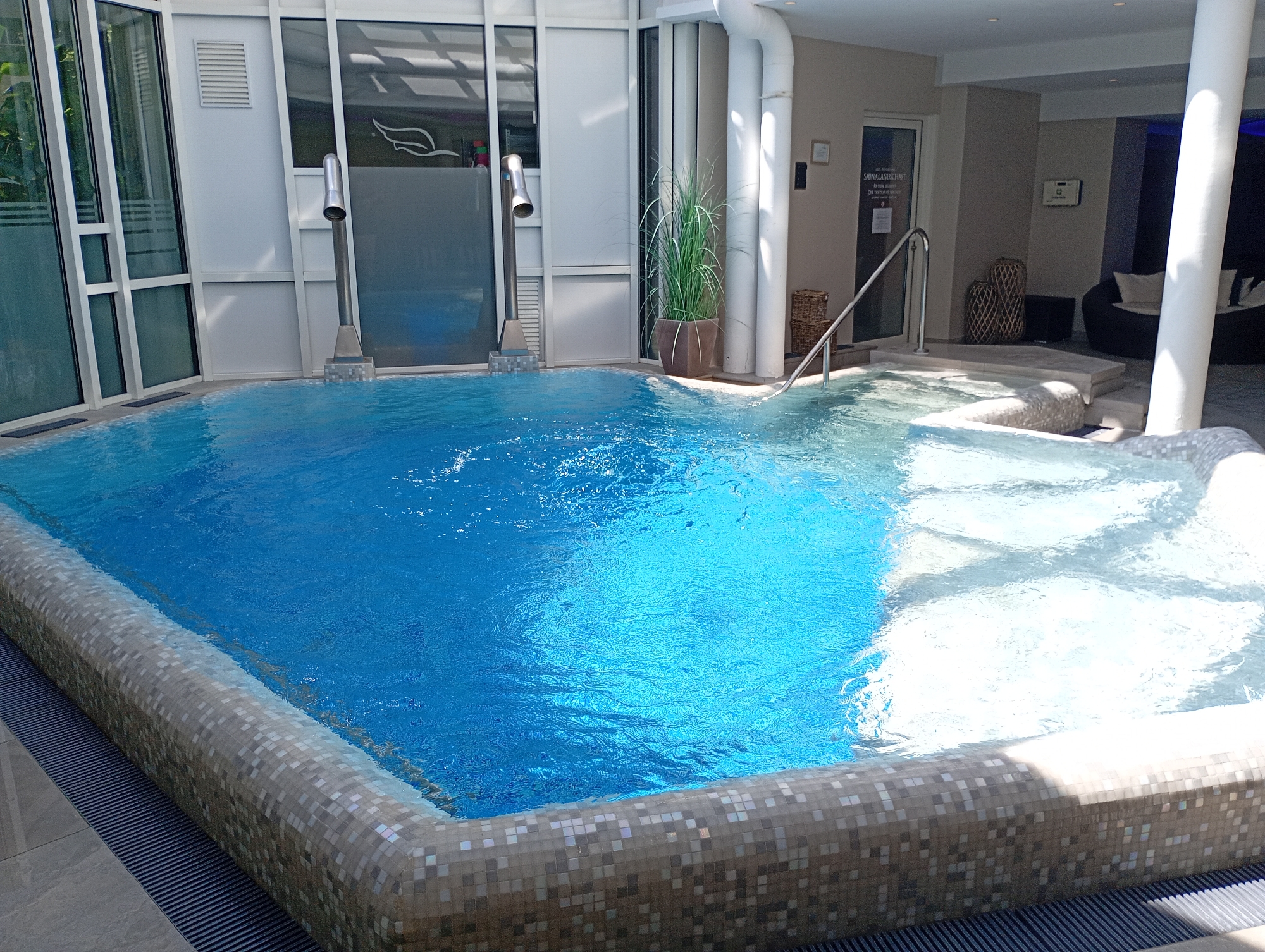 The image shows an indoor swimming pool complex in Bad Sachsa, Germany. The pool itself is rectangular and filled with clear blue water. The pool's edge is decorated with small, light-colored tiles. To the left of the pool, large glass windows allow natural light to enter the space. In the background, there are decorative elements such as potted plants, woven baskets, and lounge furniture. A metal handrail is visible, providing access into the pool. Signs on a door in the background indicate it is a spa area. The overall scene suggests a modern and relaxing spa environment.