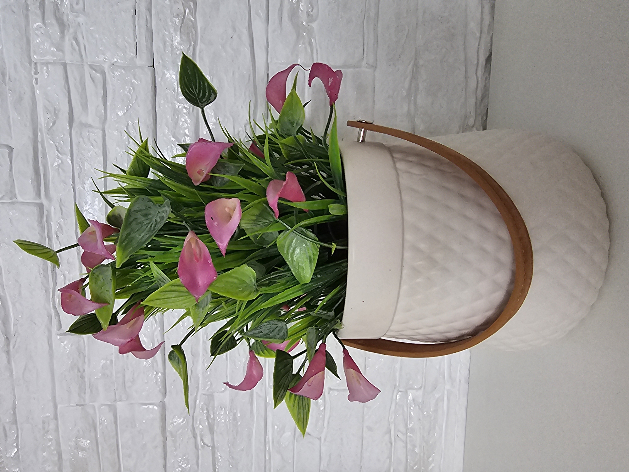An artificial potted plant with pink calla lilies and green foliage sits in a textured white ceramic pot with a brown leather handle. The pot rests on a white surface in front of a white brick-patterned wall. The scene is static and appears to be an indoor still life. There is no indication of the city of Kalinovka, Russia.