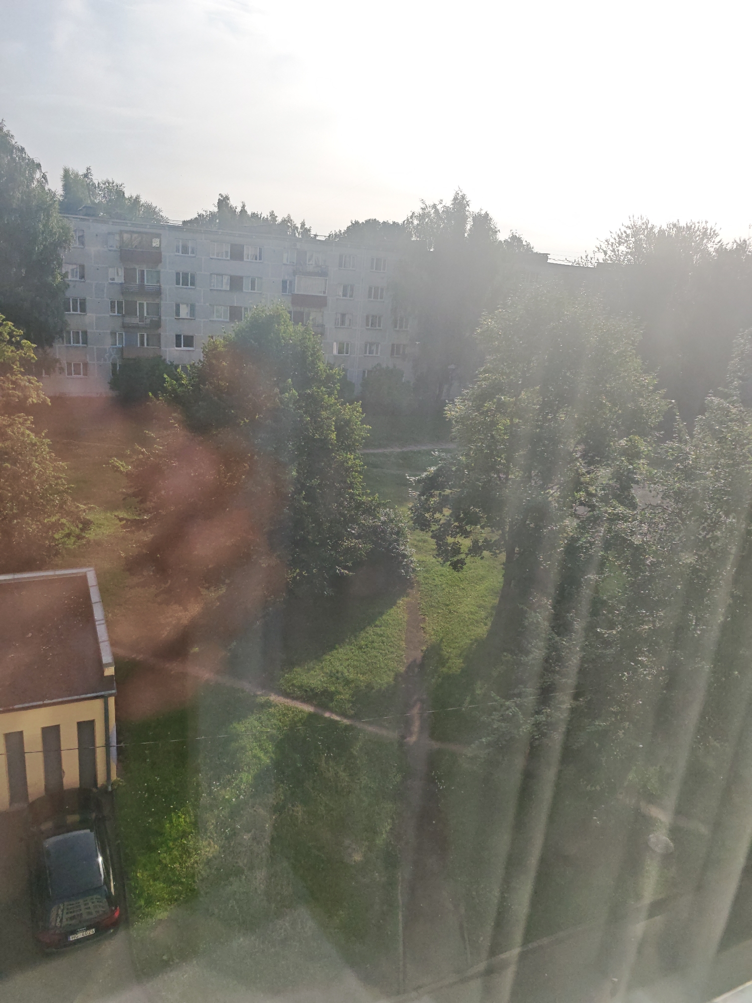 The image depicts a view from a window overlooking an apartment building and surrounding greenery in Riga, Latvia. The apartment building is a multi-story structure, likely a Soviet-era block, with numerous windows. Trees and grass are visible in the foreground and around the building, suggesting a residential area with parks or courtyards. A black car is parked next to a yellow and brown building on the left side of the frame. The sun is shining brightly, creating lens flare and glare that partially obscure parts of the image, particularly on the right side, with vertical light streaks. The overall impression is of a typical urban residential scene on a bright day.