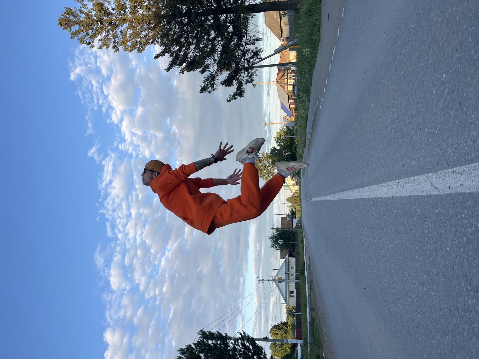 An outdoor, eye-level shot depicts a person seemingly floating horizontally above a paved road, captured in Chaikovsky, Russia. The individual, appearing male, is dressed in a bright orange hooded tracksuit, a brown cap, and white sneakers with "adidas" visible on one shoe. Tattoos are visible on his left forearm. His arms are outstretched, and knees are slightly bent, creating a dynamic pose as if suspended mid-air.
The background features a vast blue sky with scattered white cumulus clouds. Below the floating figure, a dark grey asphalt road, marked with a white dashed line, stretches into the distance, bordered by a green grassy verge on the left. Beyond the road and grass, several green trees, utility poles with overhead power lines, and residential buildings are visible, suggesting a rural or suburban setting. The lighting indicates either late afternoon or early evening, casting a soft, clear glow over the scene. The overall composition creates an illusion of weightlessness, playing with perspective and gravity.