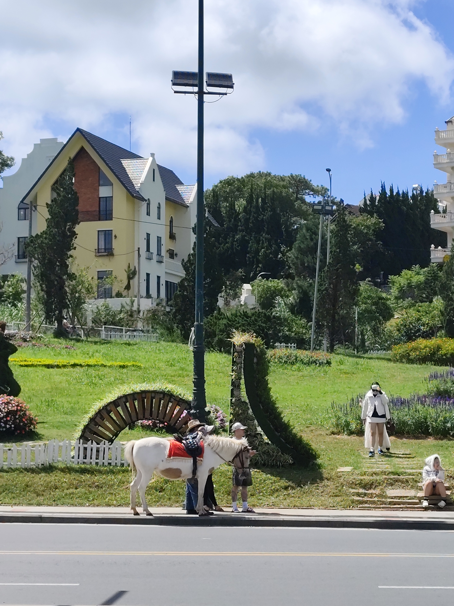 The image depicts a sunny day in Da Lat, Vietnam, likely a tourist area due to the presence of a white pony with a saddle. A young boy, wearing a hat and shorts, is holding the pony's reins. Beside him, a person in blue jeans and a dark shirt stands near the pony. In the background, a group of people are visible, including two individuals walking up a set of stairs and one person sitting on the stairs. One of the individuals standing on the stairs is wearing a white outfit and sunglasses, while the other person sitting appears to be wearing a light-colored hoodie. The environment features a lush green lawn, decorative hedges shaped into arches, and colorful flower beds. Buildings with distinct European-inspired architecture, characterized by pitched roofs and multiple stories, are visible in the background. A tall lamppost with lights stands prominently in the foreground, and a white picket fence runs along the edge of the grassy area. The scene is framed by an asphalt road in the foreground, suggesting it is a roadside attraction.