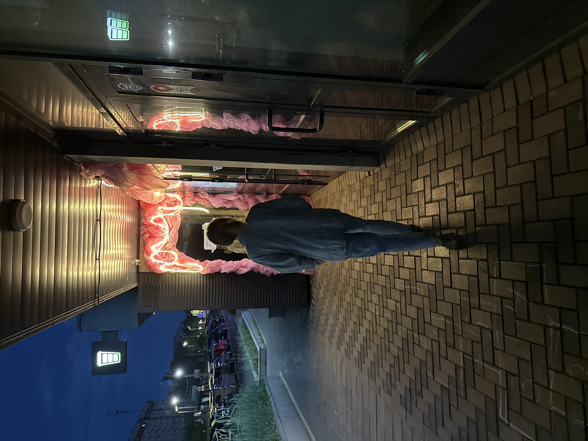 The image depicts a nighttime scene on a pedestrian walkway in Izhevsk, Russia, possibly near a cafe or shop. A person, viewed from behind, walks away from the camera down the brick-paved sidewalk. The person is wearing a light blue or grey long-sleeved shirt and blue jeans.
To the left of the walkway, there is a line of parked cars visible under streetlights, and a building with a dark facade. Above the building's entrance, a green illuminated sign shaped like a coffee cup is mounted.
To the right of the walkway, a building features large glass doors. Above the entrance, a neon light installation in a bright pink color is prominently displayed, creating a warm glow. The neon lights are shaped into decorative swirls and loops, intertwined with what appears to be pink fabric or netting. Inside the glass doors, more of the pink neon and fabric decoration is visible, suggesting an inviting and festive interior. The ground in front of the person is paved with dark brick laid in a herringbone pattern, with some white chalk markings scattered across it. The sky above is dark blue, indicating it is nighttime.
