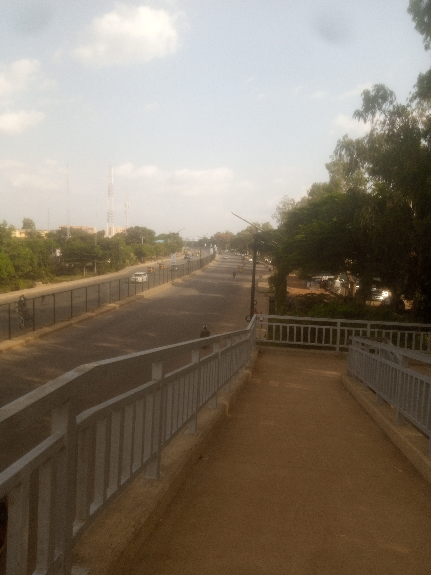 The image depicts a street scene in Unguwan Dosa, Nigeria, during the daytime. The perspective is from an elevated walkway with a concrete path and a metal railing on either side, looking down onto a road. The road appears to be a multi-lane street with a black asphalt surface. Traffic is visible on the road, including cars, motorcycles, and what appears to be a three-wheeled auto-rickshaw. Vegetation, primarily trees, lines the sides of the road, with some buildings visible in the background, suggesting an urban or semi-urban environment. Several tall communication towers are also visible in the distance. The sky is mostly clear with scattered white clouds, indicating fair weather. The lighting suggests it is likely morning or late afternoon. There is no visible text in the image.