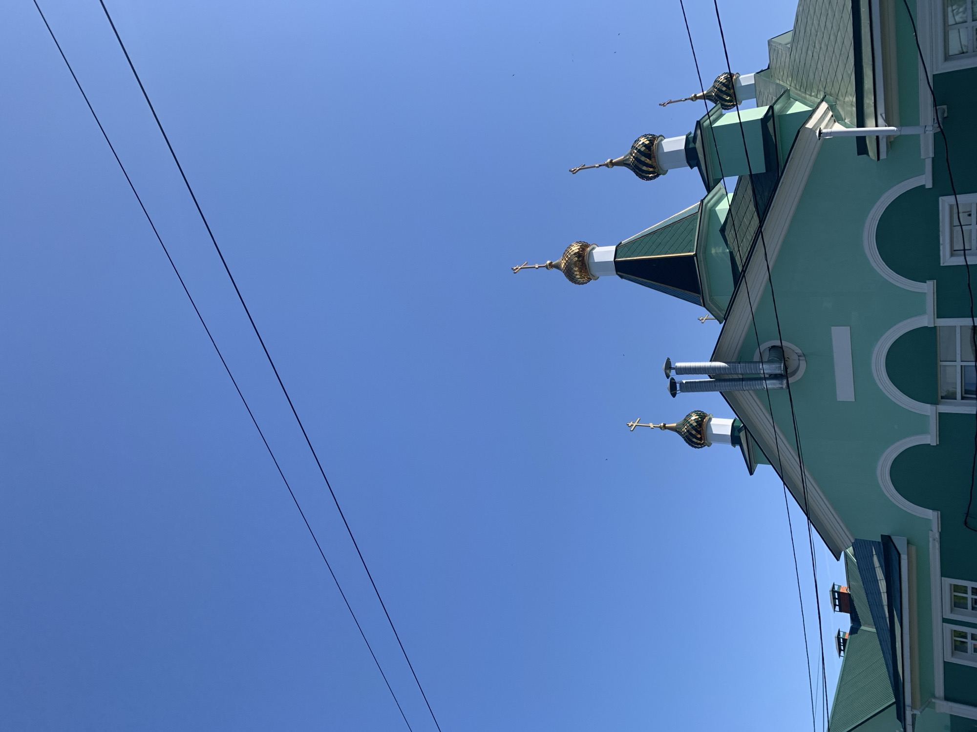 The image presents a low-angle view of a light-green building with golden onion domes and crosses, set against a clear blue sky. The building exhibits architectural features typical of Russian Orthodox churches. Three power lines stretch across the sky, diagonally from the upper-left to the lower-right of the frame. The building includes white arched details on the facade and windows with white frames. The location is likely in the city of Chaikovsky, Russia, based on the presence of Russian Orthodox architecture. No people are visible.