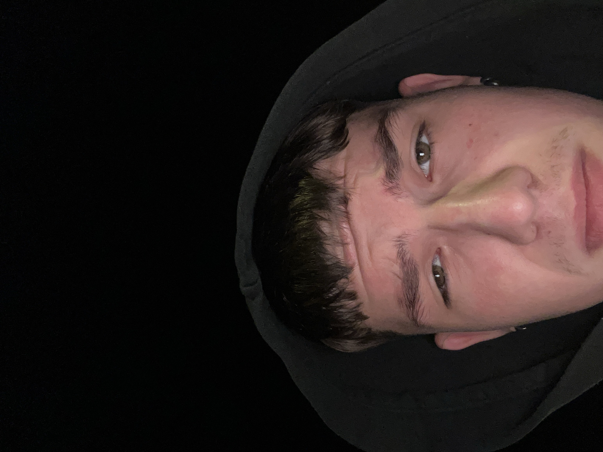 A close-up, low-angle, indoor shot shows a young person wearing a black hooded sweatshirt, their face illuminated by a light source from above and slightly to the left. The background is completely black, suggesting a dimly lit or dark environment. The person has short, dark brown hair with bangs, light brown eyes, and fair skin with some redness on the cheeks. Their eyebrows are dark and well-defined. They are wearing a small hoop earring in their visible left ear. Their facial expression appears somewhat pensive or perhaps slightly uncomfortable, with a furrowed brow and slightly downcast eyes. The image is cropped such that only the upper portion of the person's face and head are visible. The specific location in the city of Siedlce, Poland cannot be determined from the visual information provided.