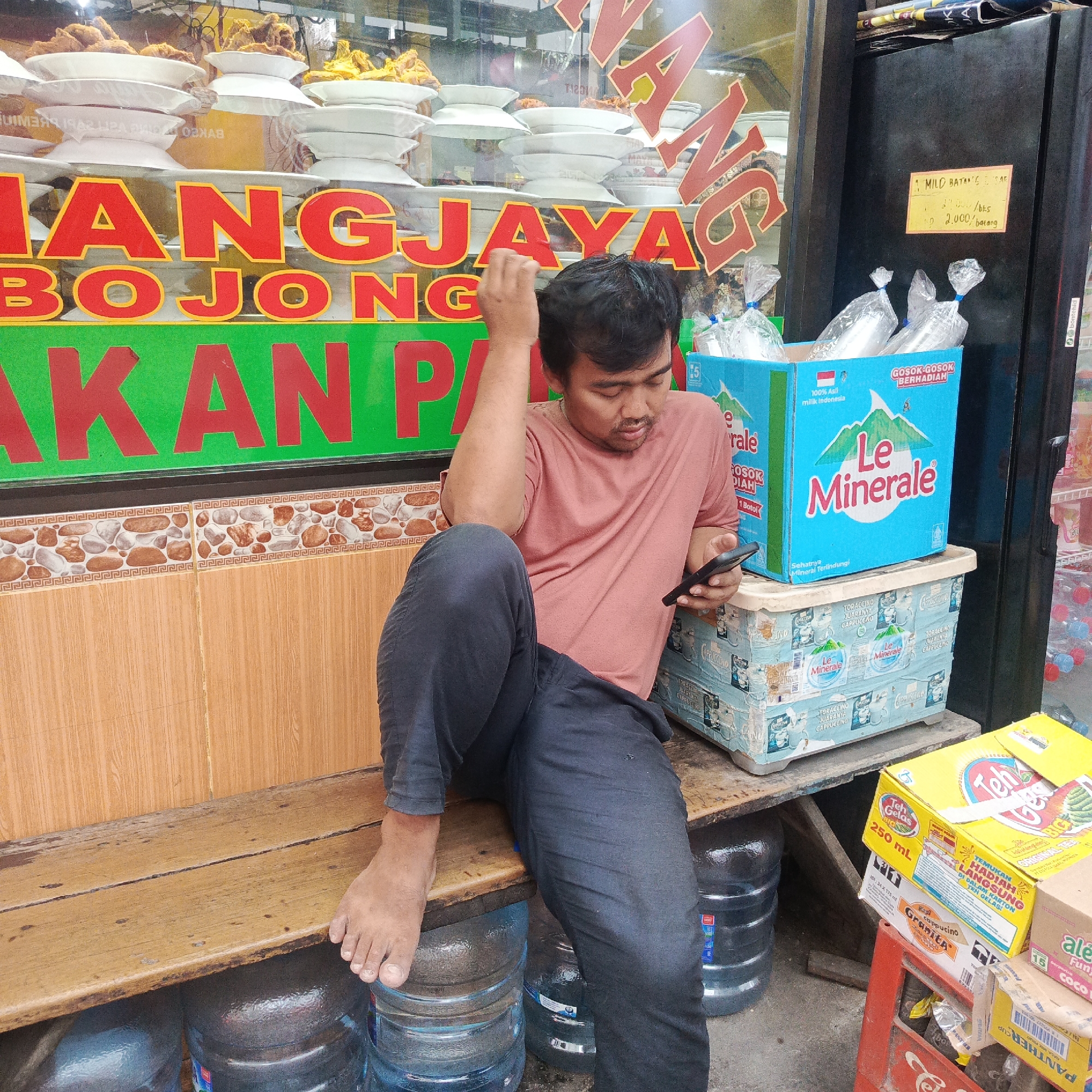 The image shows a street-side food stall in South Jakarta, Indonesia. A young man is seated on a wooden bench, looking at his mobile phone. He is wearing a coral-colored t-shirt and dark pants. Behind him, stacked cases of Le Minerale bottled water and other beverages are visible, along with large water jugs. The stall's facade has large red lettering that reads "JIANGJAYA" and "MAKAN PA" which likely indicates it is a food establishment. The overall scene suggests a casual, everyday setting within a bustling urban environment.