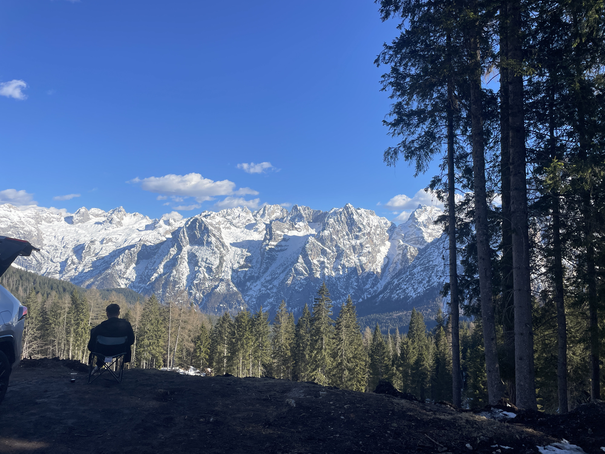 A person is sitting in a chair facing snow-capped mountains, likely in the vicinity of Cortina d'Ampezzo, Italy. The subject is positioned on a dirt clearing bordered by tall pine trees. A silver car is partially visible on the left. A cup sits on the ground near the chair. The sky is blue with scattered white clouds.