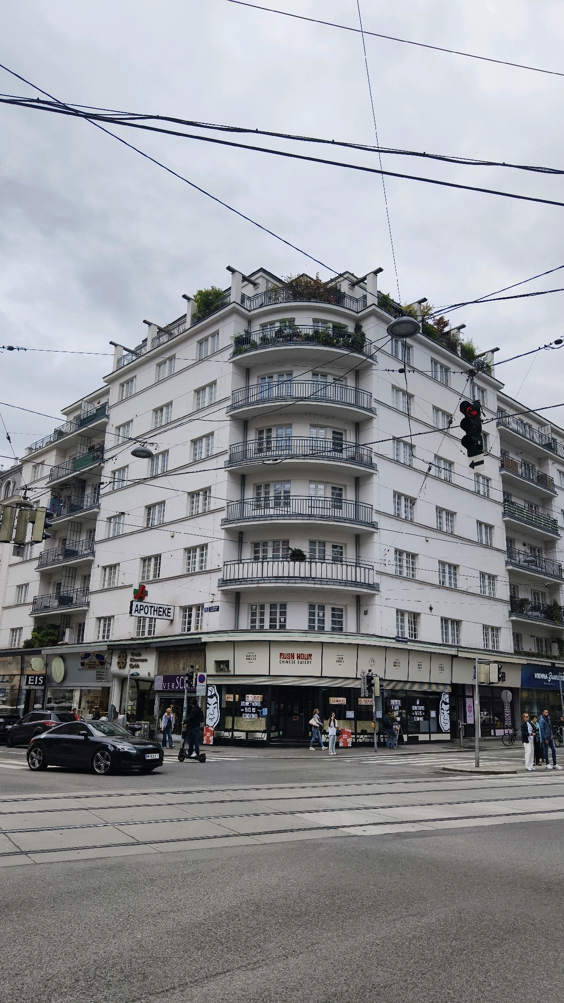 The image depicts a wide, overcast day in Vienna, Austria, focusing on a white, multi-story building with curved balconies and ornate railings. The building has a white facade with numerous windows and a rooftop garden. Multiple sets of overhead power lines crisscross the sky. Below the building, a street corner is visible with several shops, including an "APOTHEKE" (pharmacy), a Chinese eatery labeled "RUSH HOUR CHINESE EATERY," and other businesses. A traffic light is visible on the right side of the image, showing red. Several cars, including a black Audi, are on the road, and a person is riding a scooter. Tram tracks are laid across the asphalt. Pedestrians are walking on the sidewalks and crossing the street. The overall scene is a typical urban environment in Vienna.