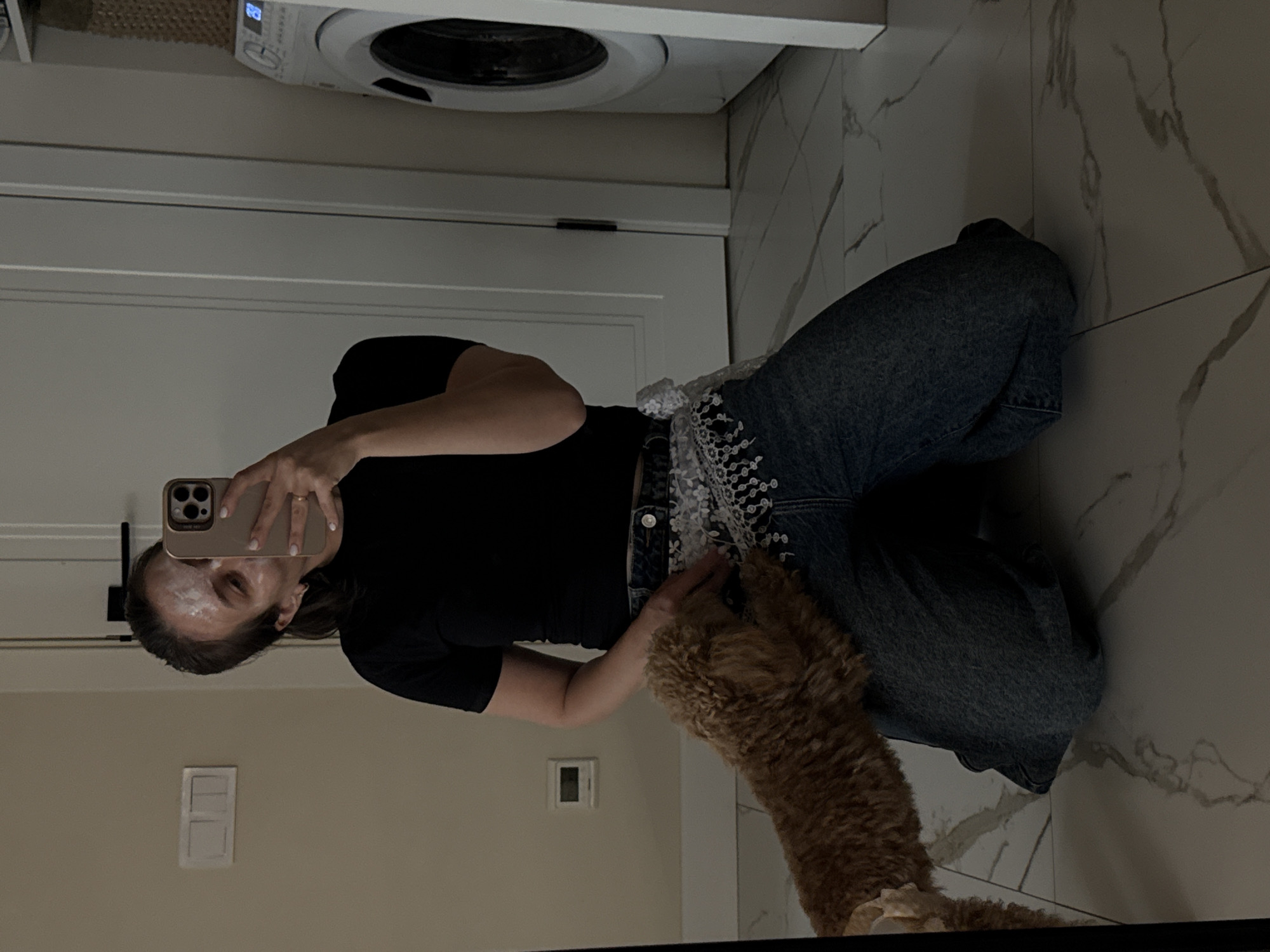 A person is depicted in a mirror selfie, kneeling on a light-colored tiled floor that features a white and gray marble pattern. The image is taken from a slightly low angle, capturing the person's reflection. The individual, appearing to be a woman, is wearing a black short-sleeved top and blue denim jeans. A notable detail on the jeans is a white, intricately embroidered or lace-like pattern along the waistline and upper thigh area. She holds a light-colored smartphone with a multi-lens camera vertically in her right hand, positioned to capture her reflection in the mirror. White residue, likely a face mask or cream, is visible on her forehead and cheeks.
Next to the person, a medium-sized dog with light brown, curly fur, resembling a goldendoodle or similar breed, is visible. The dog is positioned with its body partially obscured, appearing to lean against or interact with the person.
In the background, a white washing machine is visible atop a white countertop, with a digital display showing "28" illuminated in blue. Below the countertop, a white door with a dark handle is partially seen. The walls are light-colored, and a white light switch plate is visible in the lower-left portion of the reflection. The overall setting suggests an indoor environment, likely a utility room or bathroom in a residence located in Pervomaysky, Russia. The lighting appears to be artificial.