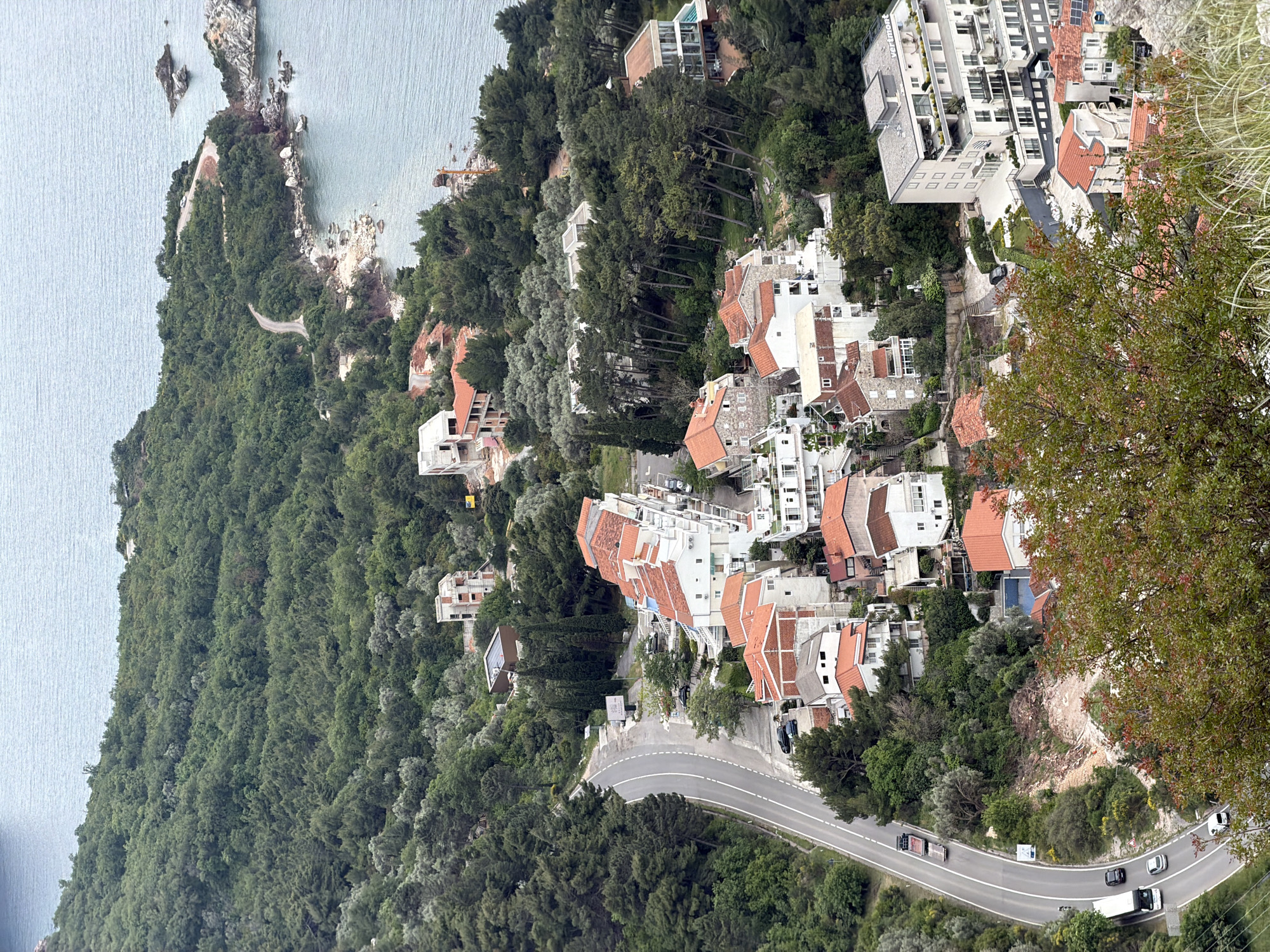 A high-angle view captures a coastal landscape near Sveti Stefan, Montenegro. The scene features dense green vegetation covering a sloped terrain, interspersed with residential buildings displaying white facades and terracotta-colored roofs. A winding asphalt road cuts through the landscape, with several cars traveling along it. A body of water, presumably the Adriatic Sea, is visible on the upper left, with a rocky coastline. The overall atmosphere is serene, depicting a mix of natural beauty and human development.