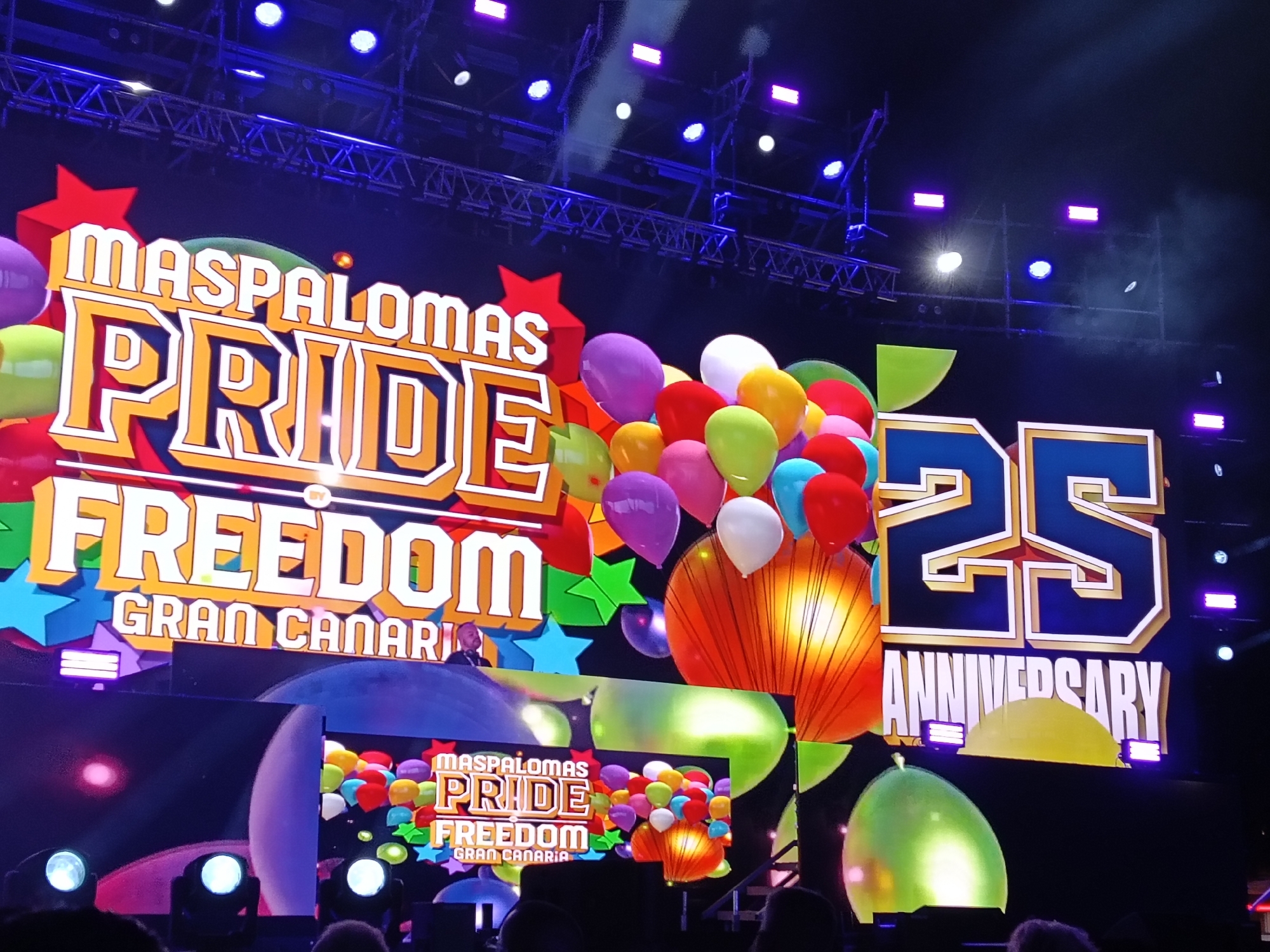 The image depicts a stage at an event celebrating the 25th anniversary of Maspalomas Pride Freedom in Gran Canaria, part of the municipality of San Bartolomé de Tirajana, Spain. The stage backdrop is a large screen displaying the event's logo, which includes the words "Maspalomas Pride Freedom Gran Canaria" in a colorful, cartoonish font. The logo is decorated with stars and balloons. A smaller screen below repeats the same image. A person stands in front of the large screen. Below the stage is an audience, whose heads are visible. The stage is lit with a variety of spotlights. The lighting rig above the stage is visible.