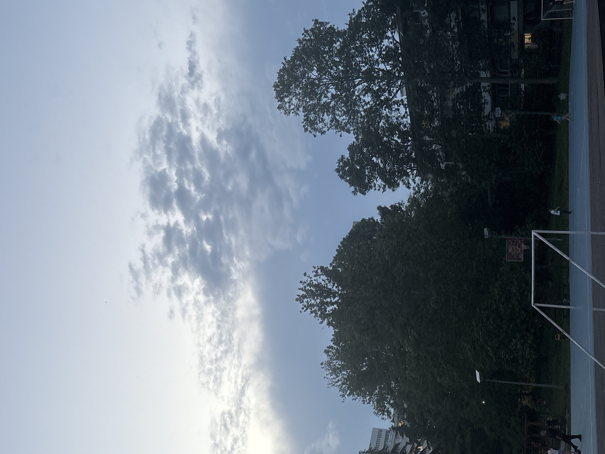 The image captures an outdoor, multi-purpose sports court in an unknown urban or suburban location during twilight or early evening. The sky displays a gradient from pale grey-blue on the upper left to scattered, dark grey clouds across the remainder of the upper frame, with a bright, diffuse light source present on the far left.
In the foreground, the court surface is light blue. A white metal soccer goal stands centrally, with its crossbar and posts clearly visible. Behind it and slightly to the right, a basketball hoop with a red backboard is situated. Two tall, dark pole-mounted streetlights with rectangular fixtures are visible on the court; one stands to the left of the basketball hoop, and another is further right.
Several individuals are present on or near the court. On the left side of the court, a person wearing dark clothing (a dark top and pants) is walking towards the right. Another person in a light blue top and shorts is visible in motion on the mid-right side of the court, suggesting activity. Other less distinct figures are visible in the background on the left side of the court.
The background is dominated by dense, dark green deciduous trees that obscure much of the surrounding architecture. Portions of multi-story buildings are visible through and above the tree canopy on both the left and right sides of the frame. Some windows in these buildings show interior illumination, consistent with the low light conditions.