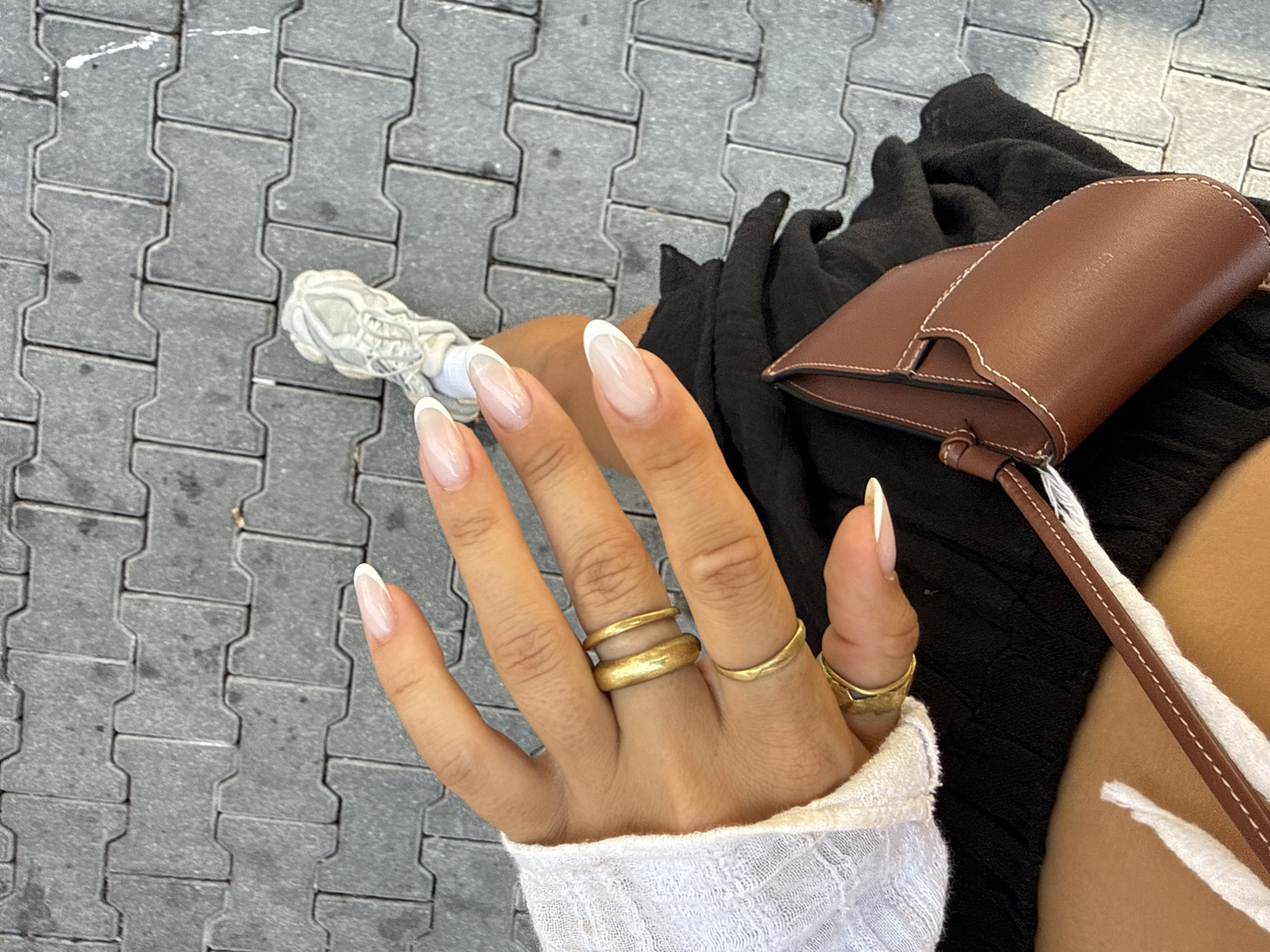 A person's hand with French manicured nails and several gold rings is held up, revealing part of a white sleeve and black clothing underneath. A small brown leather bag with white stitching is visible, resting on the black clothing. In the background, grey interlocking paving stones are seen, and a portion of a light-colored athletic shoe is present. The scene is illuminated by natural light, casting shadows. The location is the city of Fethiye, Turkey.