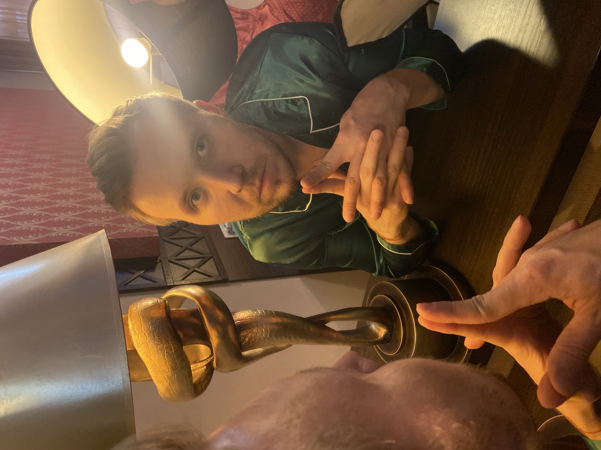 A man with short blond hair and stubble is visible, wearing a green pajama top with white piping. He is looking directly at the camera and resting his hands near his face. Next to him is a lamp with a golden base in the shape of entwined snakes. A partially visible person with fair skin and hair is close to the lamp. The scene appears to be indoors, potentially in a hotel room or private residence. The location, Yekaterinburg, Russia, cannot be confirmed from the image alone.
