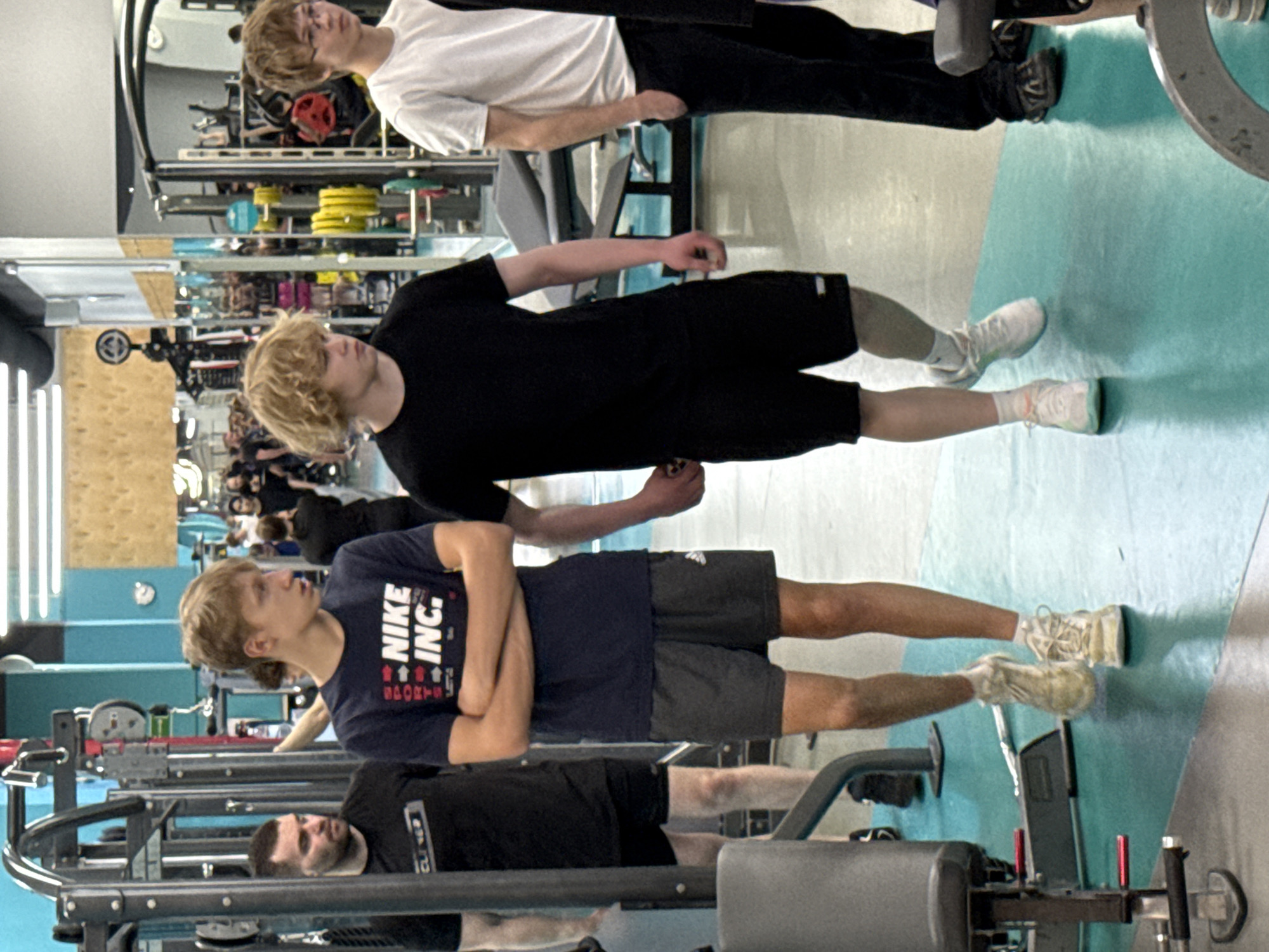 The image shows a mirrored wall in a gym. Three men are standing in the foreground and their reflections are visible in the mirror behind them. The man in the top left wears a white shirt and black pants. The second man wears a black shirt and black shorts, while the third man is wearing a dark blue Nike t-shirt and gray shorts. Weightlifting equipment is visible in front of the mirror. The specific location in Yekaterinburg, Russia, cannot be determined from the given image.