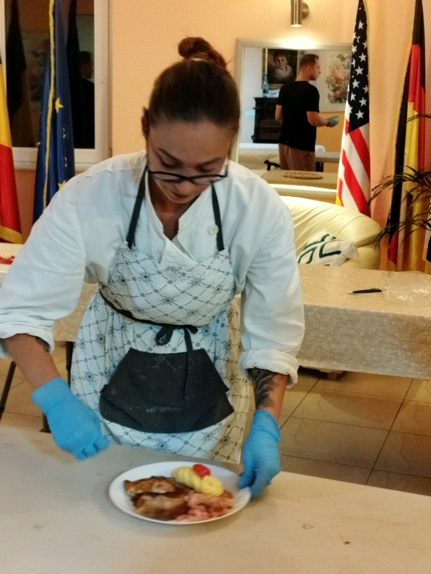 A woman wearing a white shirt and a patterned apron, with blue gloves on her hands, is presenting a plate of food. The food appears to be meat with some yellow and red garnishes. In the background, there are flags, including an American flag, and a reflection in a mirror showing a man in a black shirt and shorts. The scene appears to be indoors, with a table in the foreground and a couch and a large table in the background. The floor is tiled. The context of Bucharest, Romania is not present in the image.