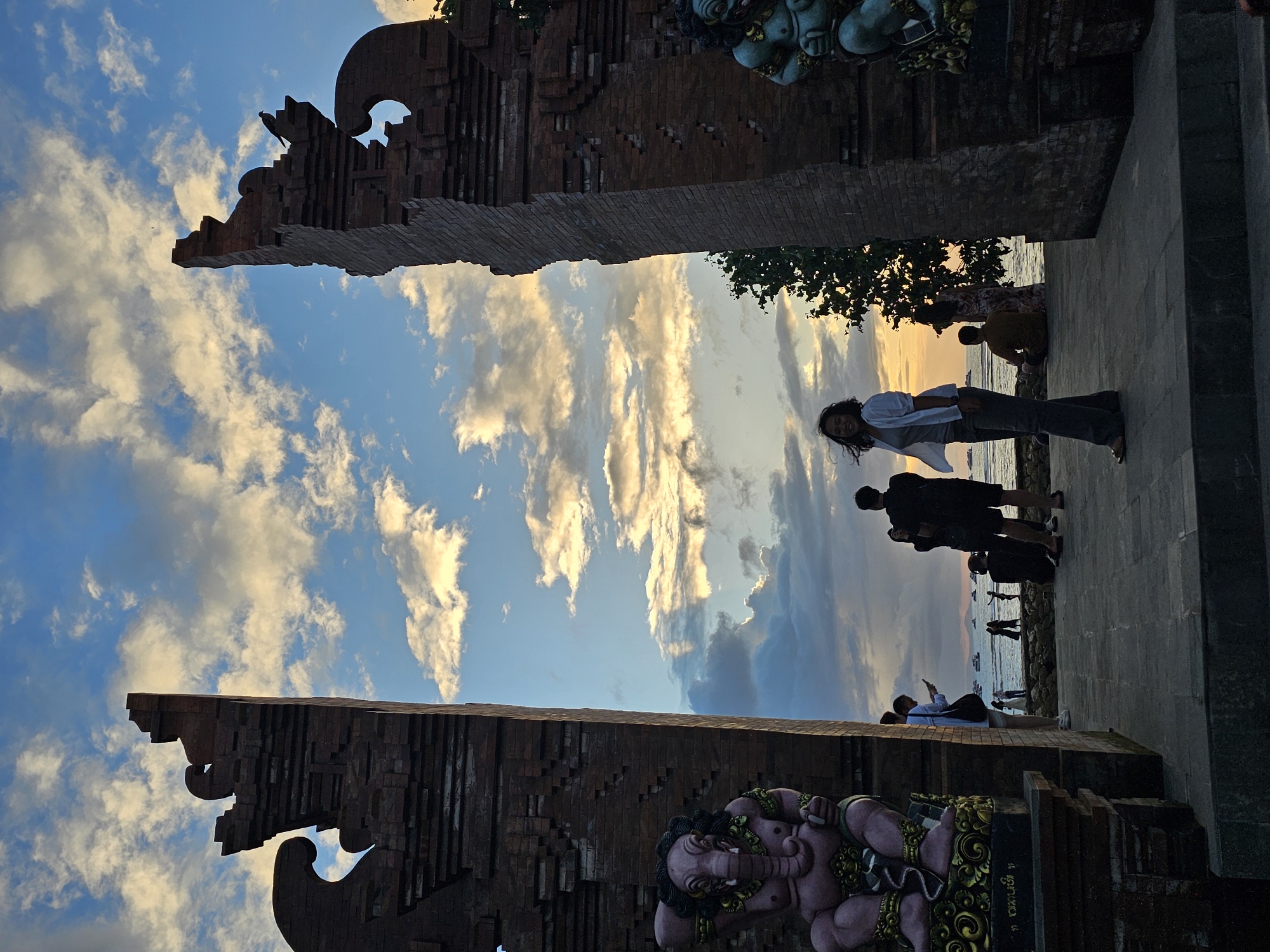 A low-angle, full-shot photograph captures a wide brick gateway adorned with two decorative statues, one of an elephant-like figure on the left and a humanoid figure on the right, against a dusky sky filled with clouds. The gateway opens to a view of the sea, where several boats are visible on the horizon. In the foreground, a woman in dark pants and a white shirt is standing, facing the camera with a slight smile. Other people are scattered around the scene, some near the water, others near the gateway. The overall ambiance is that of a sunset gathering at a coastal location. This scene is located at the north entrance of the Bajra Sandhi Monument in Denpasar, Indonesia, a landmark commemorating Balinese people's struggle against colonial rule.
