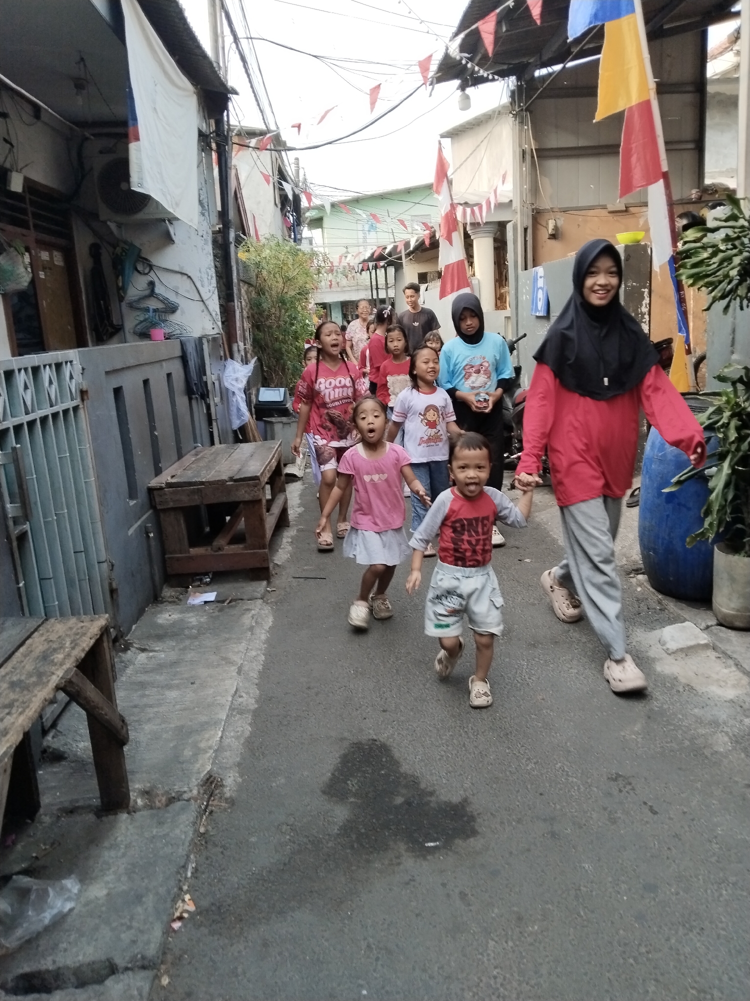 In North Jakarta, Indonesia, a group of children and one adult are walking down a narrow, paved alleyway between residential buildings. The adult, wearing a black hijab and a red top with grey pants, is leading the procession, holding the hand of a young boy in a red and grey t-shirt and shorts. The boy is looking forward with an open mouth. Behind them, several other children, mostly girls in pink and light-colored shirts, are following. Some of the children are looking towards the camera. The buildings on either side of the alley are modest, with visible wiring and air conditioning units. Red and white pennants and larger red and white flags are strung across the alley overhead, suggesting a celebration or national holiday. A large blue cylindrical object, possibly a water tank, is visible on the right side of the alley, along with potted plants. Wooden benches and tables are placed along the left side of the alley. The ground is damp in places.