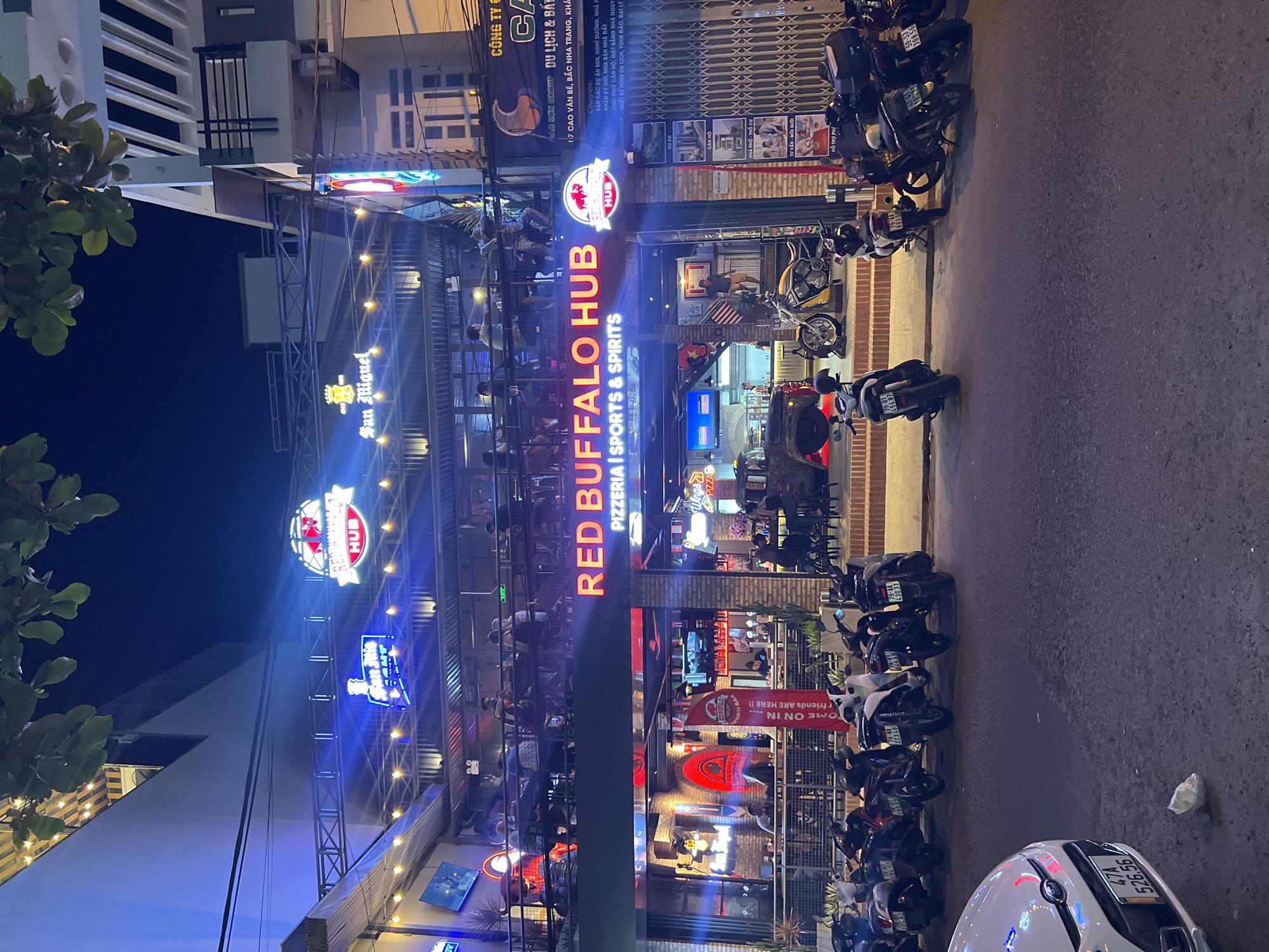 Here is a concise description of the image:
The image shows the exterior of Red Buffalo Hub, a pizzeria, sports bar and spirits establishment, at night in Bắc Nha Trang, Vietnam. The building has an open-air, multi-tiered seating area with visible patrons. Neon signs for Red Buffalo Hub, San Miguel, and other advertisements are lit. A row of parked motorbikes lines the street in front of the building. The building is on 117 Cao Văn Bé Street in Bắc Nha Trang.