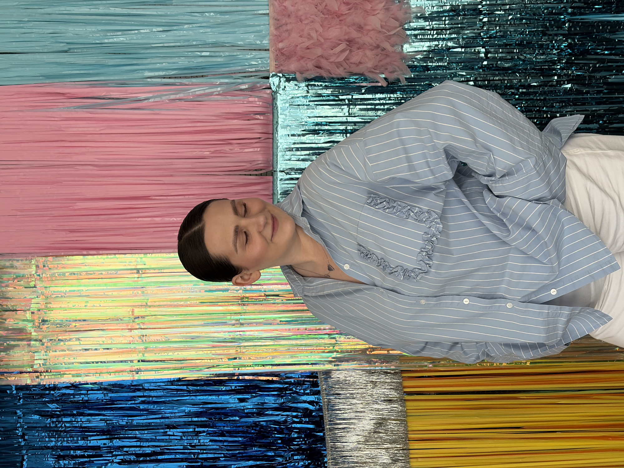A woman stands in front of a colorful backdrop composed of vertical fringe curtains in various colors, including blue, yellow, pink, and teal. She is wearing a light blue and white striped button-down shirt with ruffles on the pocket and white pants. Her eyes are closed, and she is looking down and to her right, with a slight smile. Her hair is pulled back in a bun. The overall impression is of a playful and festive atmosphere, possibly at a party or event. The location within Бараўлянскі сельскі Савет, Belarus is not discernible from the image.