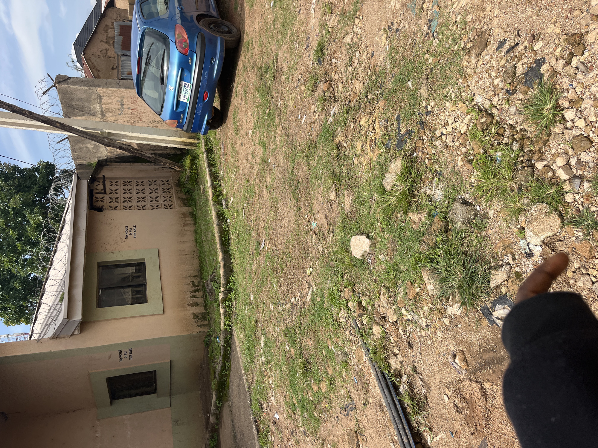 The image depicts an exterior view of a building and its surroundings in Jos, Nigeria. The building, a single-story structure, has a light brown facade with a green trim around its windows and a light gray roof. Barbed wire is visible along the top of the wall and roofline, suggesting security measures. Two windows are visible on the front of the building, and text is painted on the walls near them that reads "THIS HOUSE IS NOT FOR SALE." A blue Peugeot 206 car is parked to the right of the building. The ground in front of the building is unpaved, with patches of dry grass, dirt, and scattered rocks. Several black pipes are lying on the ground in the foreground. In the lower right corner of the image, a person's hand and arm are visible, with the hand pointing towards the ground. The sky is partly cloudy.