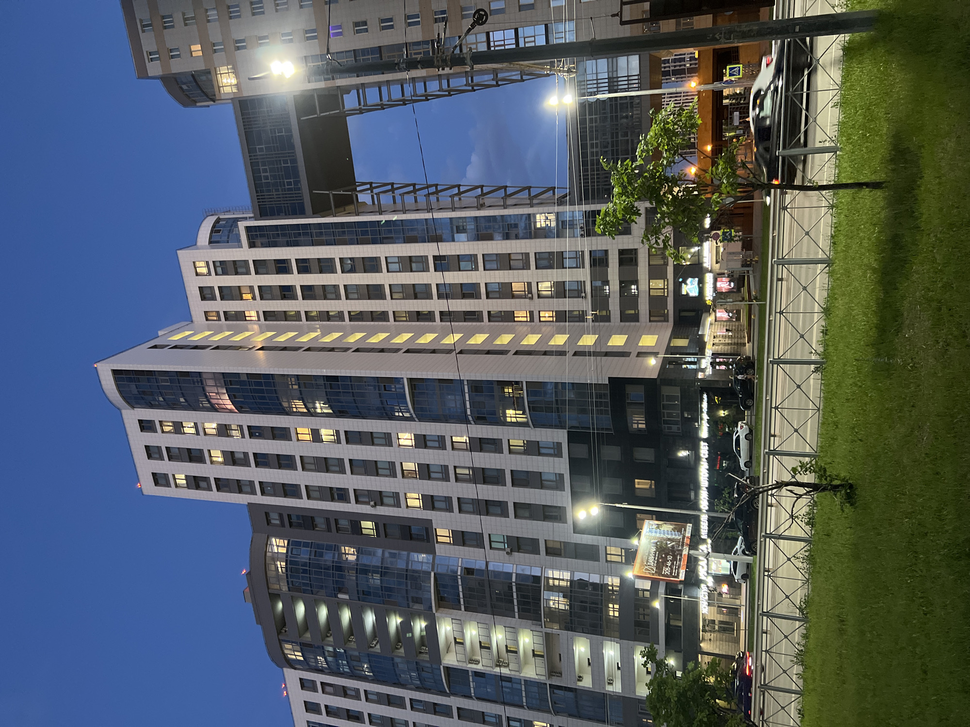 The image captures a modern apartment complex in the city of Dachnoe, Russia, at twilight. The multi-story buildings, characterized by a combination of white panels and dark blue glass facades, are illuminated by numerous yellow lights visible through the windows. A prominent architectural feature is a skybridge connecting two sections of the buildings, creating an open, arch-like structure against the darkening blue sky. Streetlights are visible, casting a bright glow, and a utility pole with wires stands to the right of the frame. Below the buildings, a street is active with car traffic, including a dark sedan moving past. A metal fence separates the street from a grassy area in the foreground, where a young tree stands. A billboard with text in Russian is mounted on the side of one of the buildings.